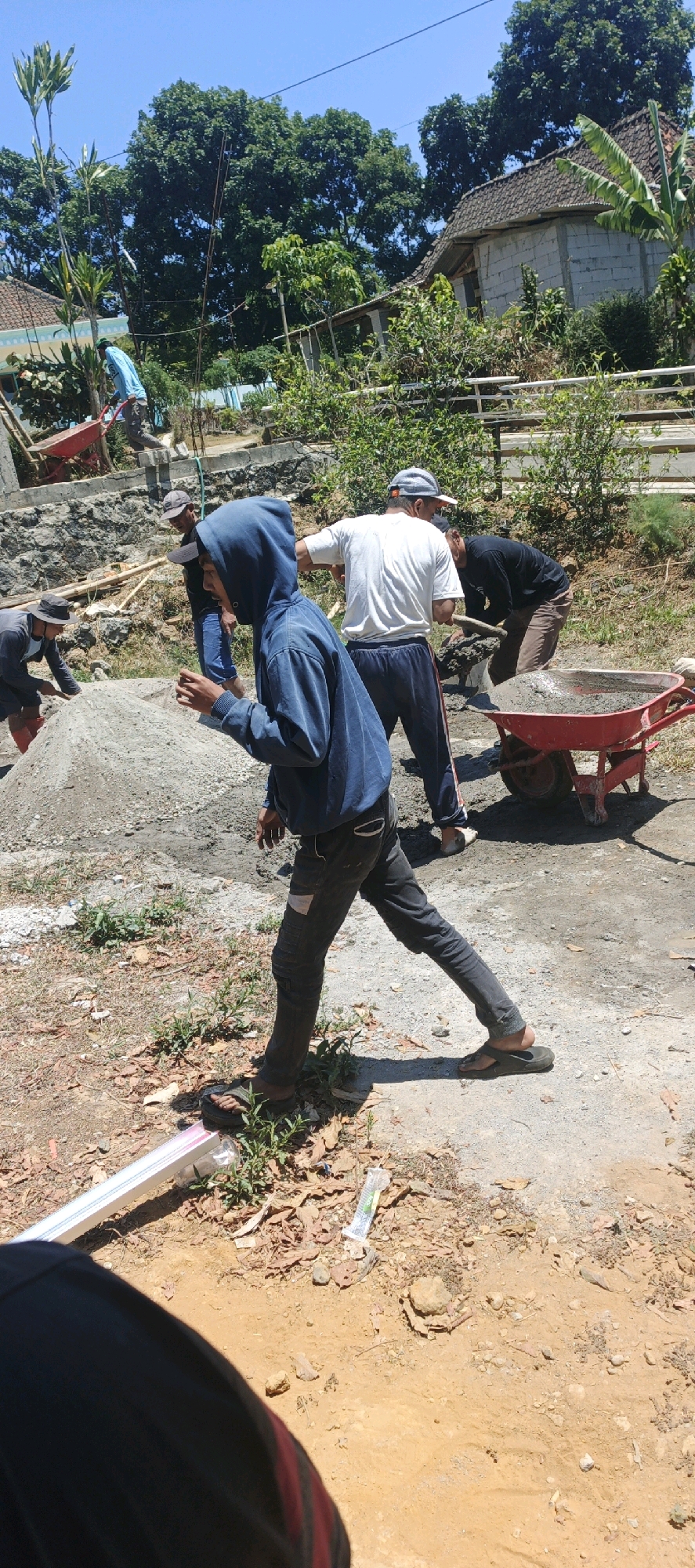 The image depicts an outdoor construction scene, likely in Jlarem, Indonesia, on a clear day. Several individuals are engaged in construction work. In the foreground, a person wearing a blue hooded sweatshirt and dark pants is captured mid-stride, with their left leg extended and their right leg bent. This individual is wearing dark sandals. Behind them, another person in a white t-shirt and dark pants is visible. To the right, a red wheelbarrow filled with what appears to be cement or a similar construction material is present. Further in the background, more people are seen working, including one individual shoveling material. The environment consists of dry, dirt ground with scattered debris, piles of construction materials like gravel, and some vegetation. In the background, houses with tiled roofs and trees can be observed, suggesting a semi-rural or residential area. A light blue sky dominates the upper portion of the image. The overall impression is one of active manual labor in an outdoor setting.