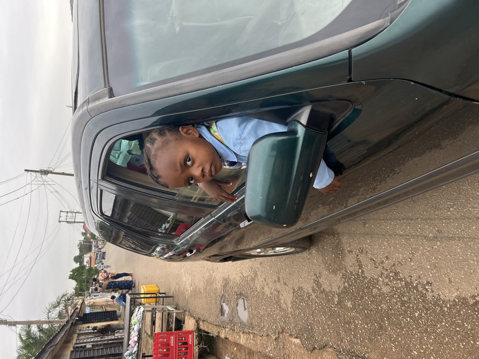 A child looks out of the open window of a dark green car. The vehicle is parked on a street with a rough, patchy surface. In the background, there are buildings and other people, suggesting an urban setting in Jos, Nigeria. A red crate marked "Coca-Cola" is near a gutter. Overhead power lines are visible. The sky is overcast.