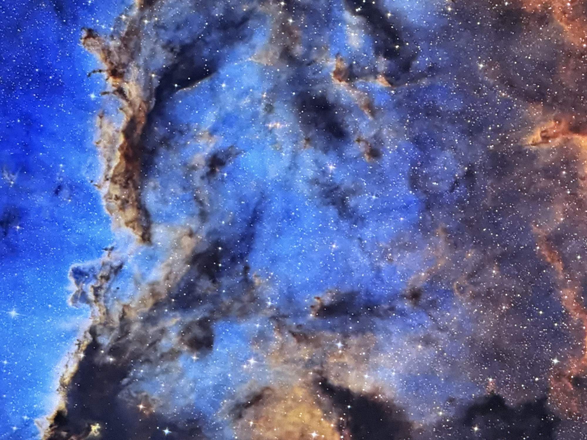 This is a close-up, detailed image of a nebula in space. The dominant colors are vibrant blues, deep purples, and fiery oranges and browns, characteristic of ionized gases and dust clouds. Countless small, bright white stars are scattered throughout the image, some appearing as sharp pinpricks of light, while others are softened by the surrounding gas. Dark, irregular shapes of dust obscure portions of the nebula, creating a sense of depth and texture. The composition is abstract, focusing on the swirling patterns and glowing luminescence of the celestial phenomenon. There are no discernible landmarks, buildings, or any indicators of a terrestrial location.