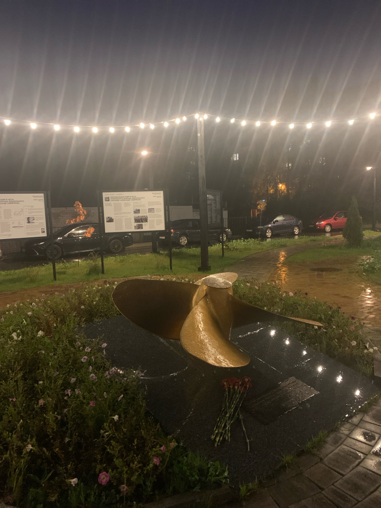 The image depicts a nighttime scene in Arkhangelsk, Russia, likely a park or public square. A large, golden ship's propeller is prominently displayed on a dark, wet stone base, adorned with a small bouquet of red flowers. The ground appears wet, suggesting recent rain, and a cobblestone path reflects the light. String lights illuminate the scene from above, creating a festive but slightly eerie atmosphere. In the background, several cars are parked, and illuminated billboards or information stands are visible. The overall impression is one of a memorial or historical site, perhaps dedicated to maritime history, given the presence of the propeller and the informational displays.