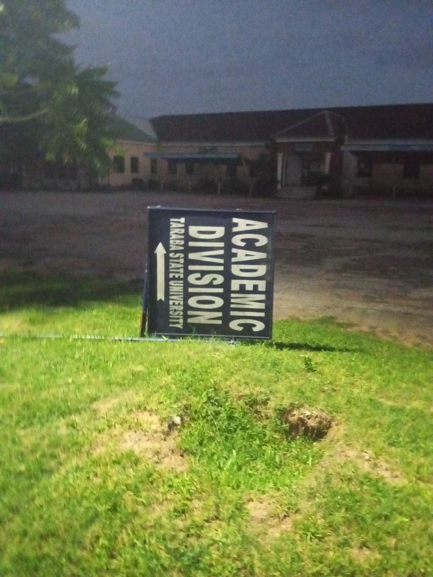 The image depicts a signpost indicating "ACADEMIC DIVISION" and "TARABA STATE UNIVERSITY" with an arrow pointing left. The signpost is positioned in a grassy area, with a building visible in the background. The lighting suggests it is either dusk or nighttime, with artificial lights illuminating the scene. The environment appears to be an outdoor space, possibly a campus or public area within Jalingo, Nigeria, given the mention of Taraba State University, which is located there. There are no people visible in the image. The overall scene is calm and lacks any dynamic activity.