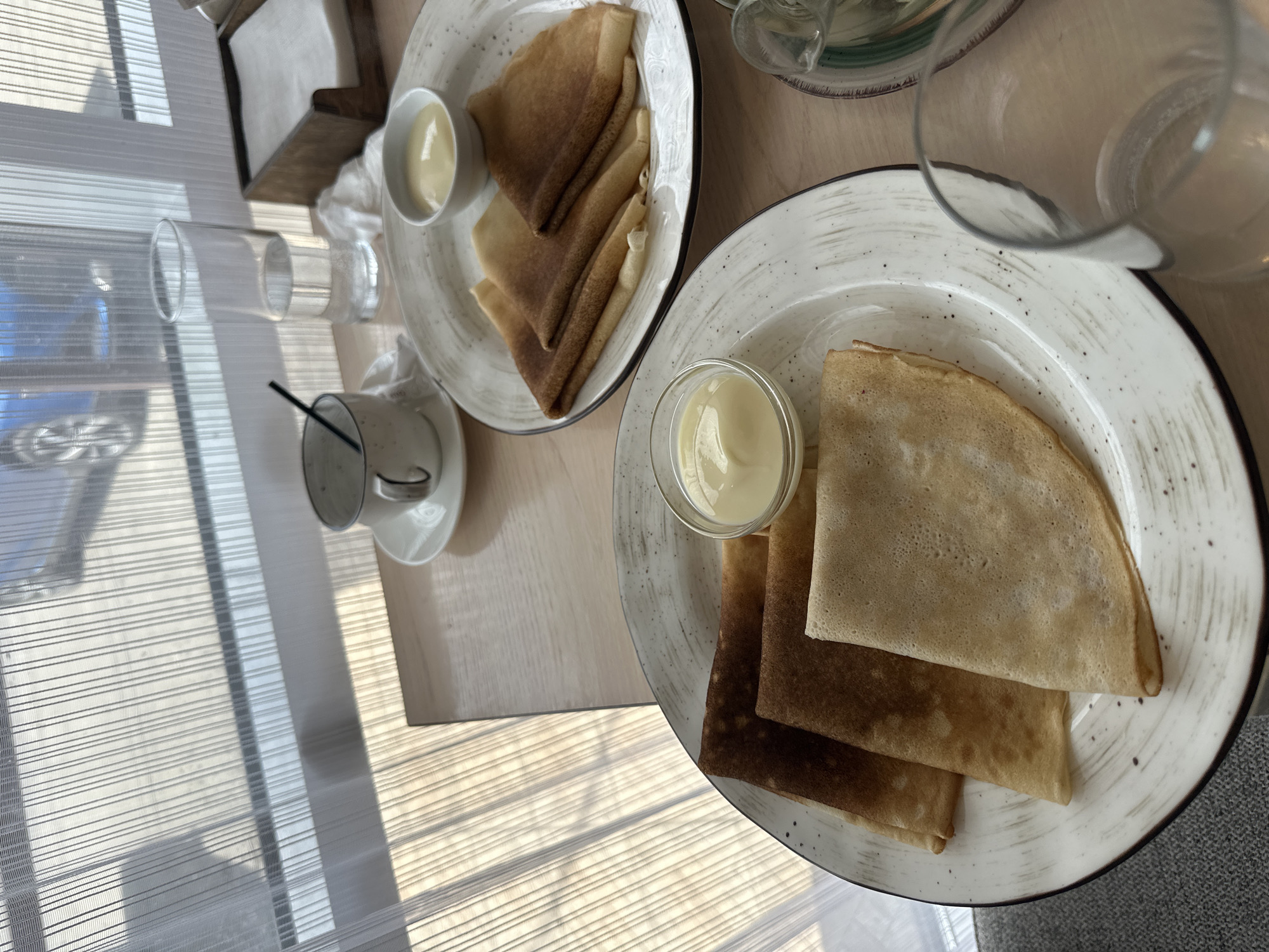 The image displays two plates of crepes with bowls of custard on a wooden table, set against a backdrop of sheer white curtains. The curtains are illuminated by sunlight, casting striped shadows on the table and the floor, and a faint reflection of a blue car is visible through them. In the background, a glass of water, a cup and saucer with a straw, and a napkin holder are also present. The scene suggests a meal or snack being enjoyed indoors, possibly at a cafe or restaurant. The location is identified as Slyudyanka, Russia.