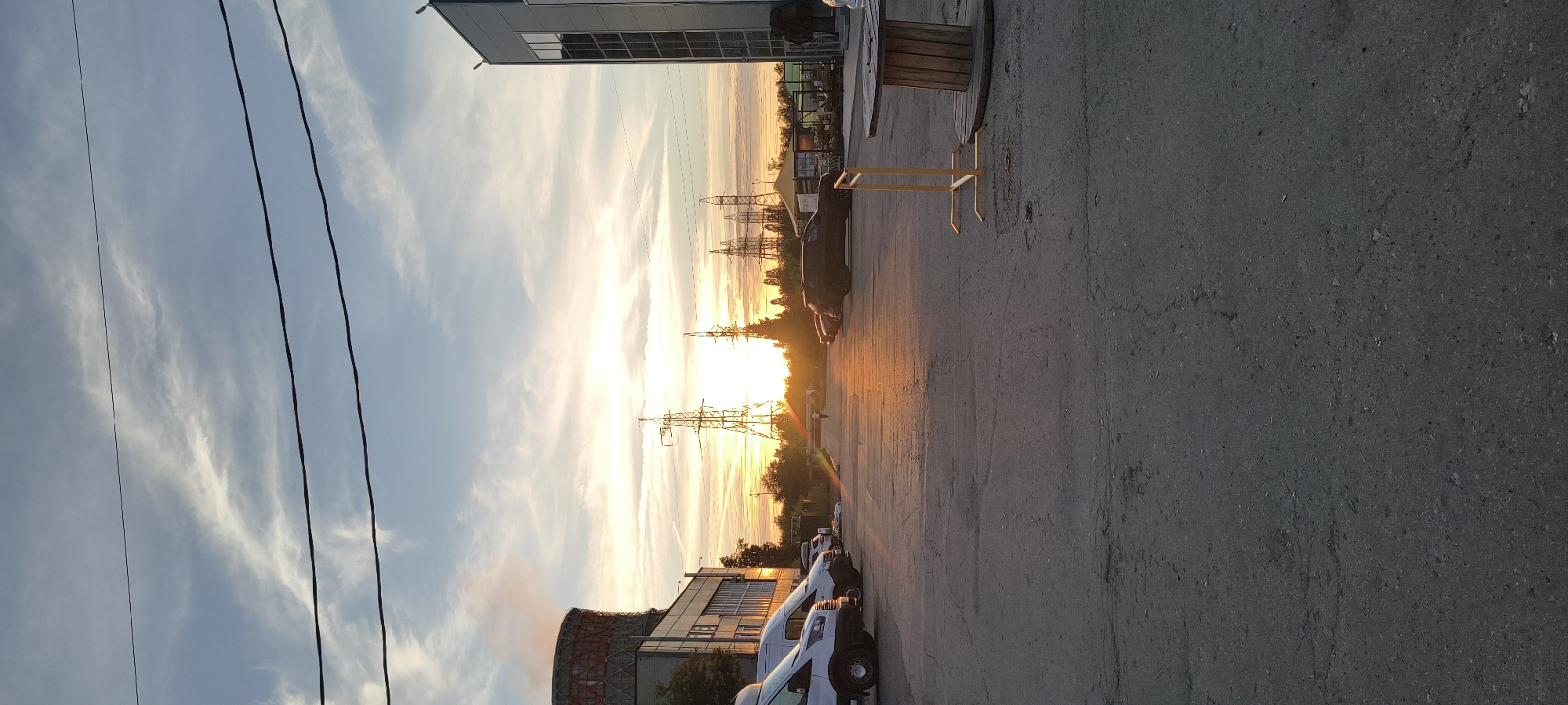 The image depicts a sunset scene in what appears to be an industrial or commercial area. The sky is filled with wispy clouds and a vibrant orange glow from the setting sun, which is positioned behind several tall electrical transmission towers. In the foreground, a paved surface, likely a parking lot or road, is visible with cracks and an uneven texture. Several vehicles are parked on the left side of the image, including white vans and a black SUV. A large industrial building with a cylindrical structure emitting smoke or steam is on the left, and another modern building with reflective windows is on the right. A large wooden spool and a yellow metal stand are also present in the foreground. A single person is walking in the distance towards the sunset. Power lines are visible stretching across the top of the frame.