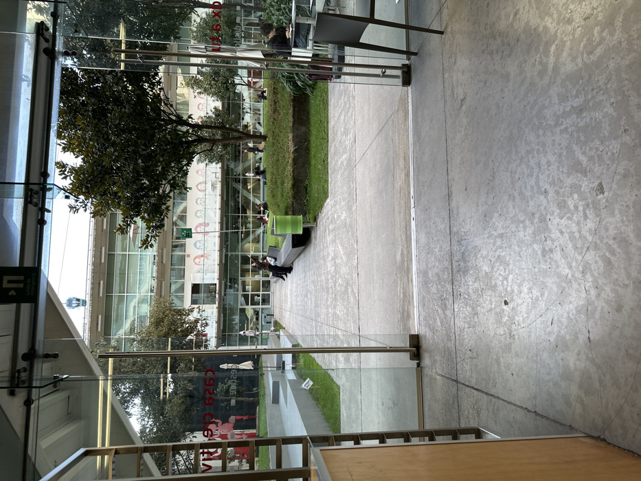 Through glass doors, a concrete walkway leads to a green space. People are sitting on benches. A large, modern building with glass walls stands in the background, partly obscured by trees. Reflections and texts are visible on the glass. The concrete floor inside is gray and rough.