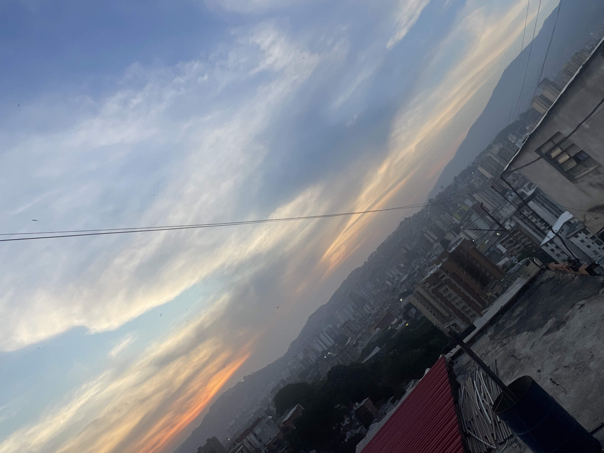 The image shows a cityscape at dusk, likely Caracas, Venezuela, based on the buildings, terrain, and context. The view is from a high vantage point, possibly a rooftop. The sky has a combination of blue and orange hues, indicating sunset or sunrise. Clouds are present. A mountain range forms the backdrop of the city. The buildings are densely packed, showing a mix of low-rise and high-rise structures. A building is in the immediate foreground, with what appears to be a corrugated metal roof and other construction materials. There is a metal pole extending from the rooftop. Several birds are in the sky. Utility wires stretch across the scene.