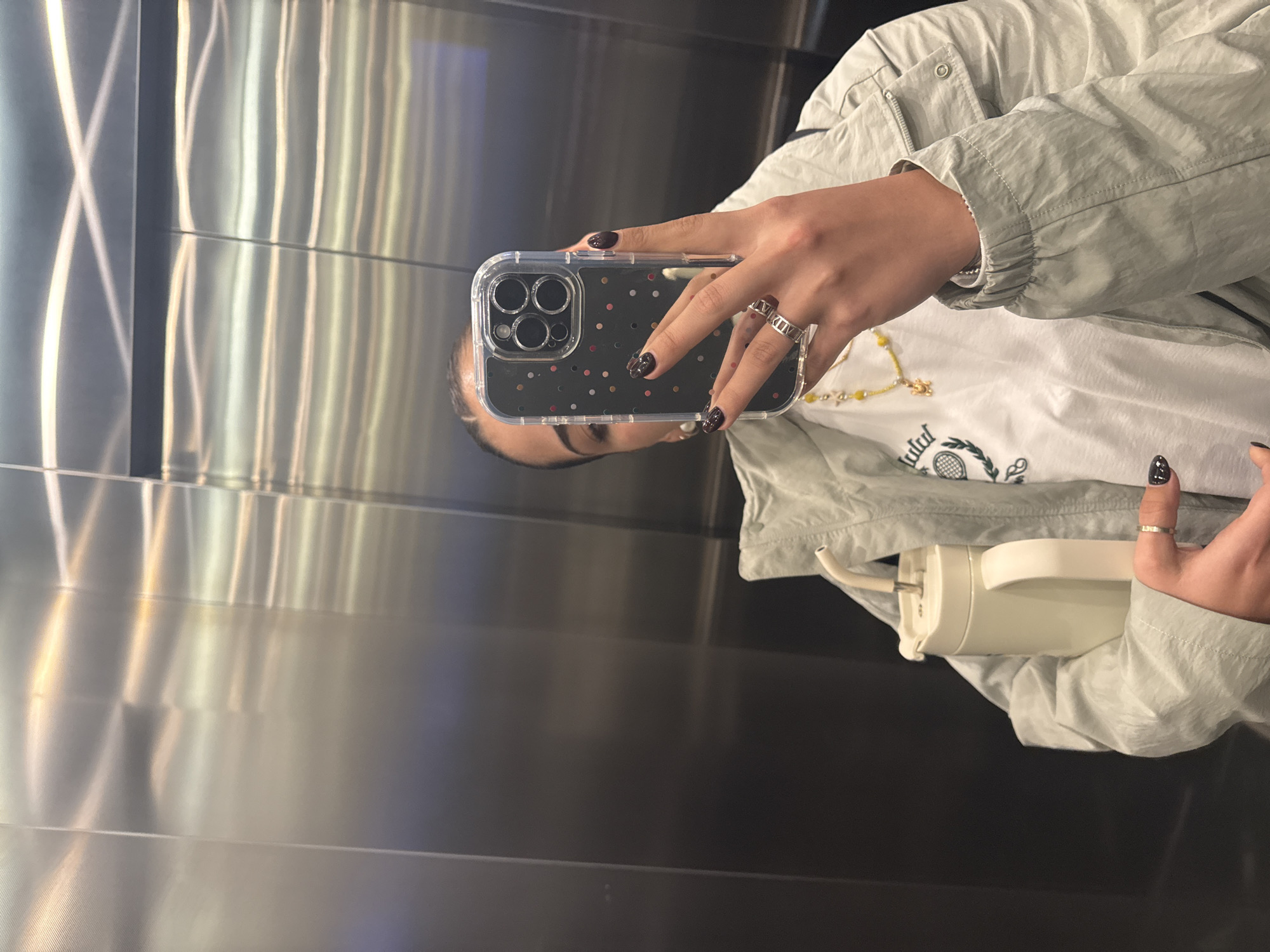 A person is taking a selfie in what appears to be an elevator. The elevator has stainless steel walls with vertical grooves, and is likely located in the city of Puebla, Mexico. The person is holding a smartphone with a clear case covered in confetti dot pattern, which obscures part of their face. The individual is wearing a light gray jacket, and a white t-shirt. The person wears multiple rings on her fingers and has painted her fingernails dark with a white design. In their hand, they hold a portable electric water flosser.