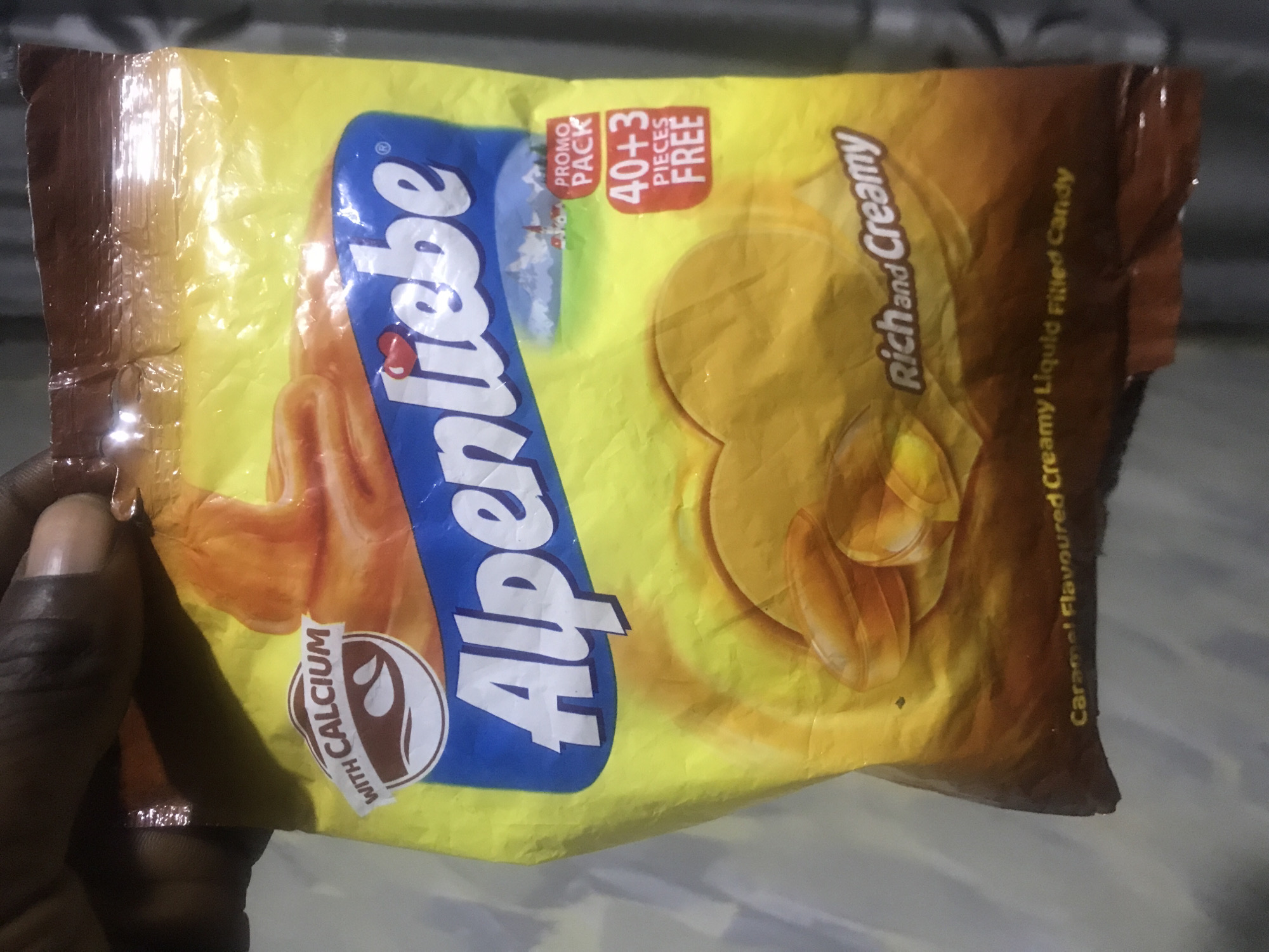 The image displays a close-up of a hand holding a yellow and brown package of Alpenliebe candy. The packaging is bright yellow with a brown border on the top and bottom edges, suggesting a sealed candy bag. The primary subject is the Alpenliebe candy wrapper, which features the brand name "Alpenliebe" in a distinctive blue and white font running diagonally across the center.
Notable text details on the wrapper include "WITH CALCIUM" printed on a circular emblem in the lower-left corner. In the upper right corner, a red box indicates a "PROMO PACK" offering "40+3 PIECES FREE." The phrase "Rich and Creamy" is written in a stylized font, and the product is further described as "Caramel Flavoured Creamy Liquid Filled Candy." Visual elements on the wrapper depict several individual caramel candies and what appears to be flowing caramel.
The hand holding the package is visible on the left side, showing dark skin and a manicured thumb. The background is out of focus, appearing to be a light-colored, possibly fabric, surface. The lighting suggests an indoor setting, and no specific time of day or weather conditions can be determined from the image. The image captures the candy packaging in detail, highlighting its promotional aspects and flavor profile.