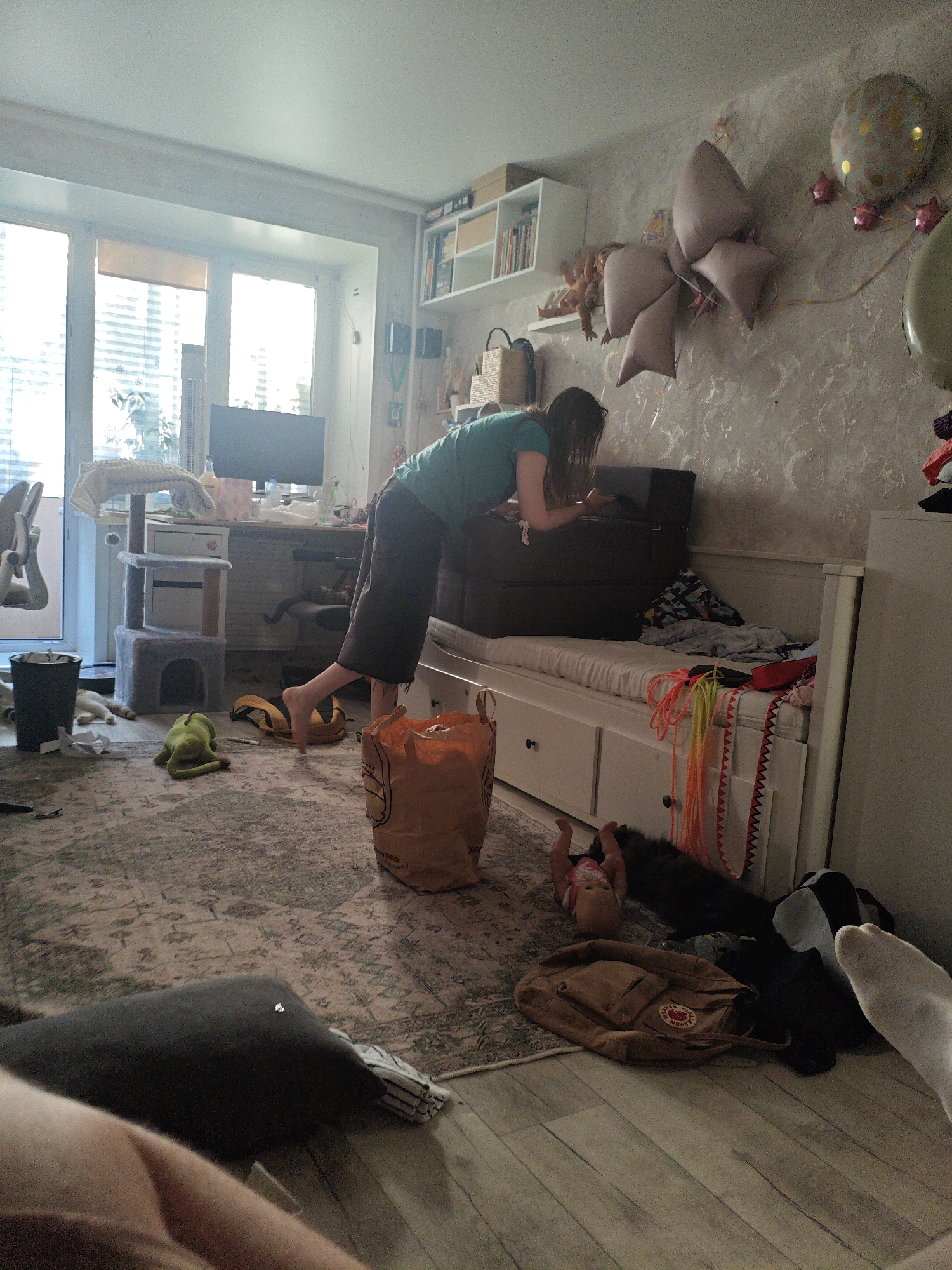The image depicts a cluttered indoor scene. A woman in a green t-shirt and dark pants is leaning over two dark brown storage containers stacked on a white daybed. The daybed is covered with a white sheet and various items of clothing. To the left of the daybed, a grey cat tree with two levels is visible, with a white blanket draped over the top level. A black trash can is located to the left of the cat tree, with crumpled white paper scattered on the floor. A green plush frog toy lies on the patterned rug in front of the cat tree. A large orange paper bag is situated to the right of the frog toy. Further to the right, a small doll lies on its back near a brown backpack. The room features a window with blinds and a desk with a computer monitor. Balloons and decorative fabric bows are attached to the wall in the background. The floor is a mix of patterned rug and light-colored wood planks. The overall environment appears to be a bedroom or living area that is in disarray. There is no information in the image that allows for the determination of the location in the city of Berdsk municipality, Russia.