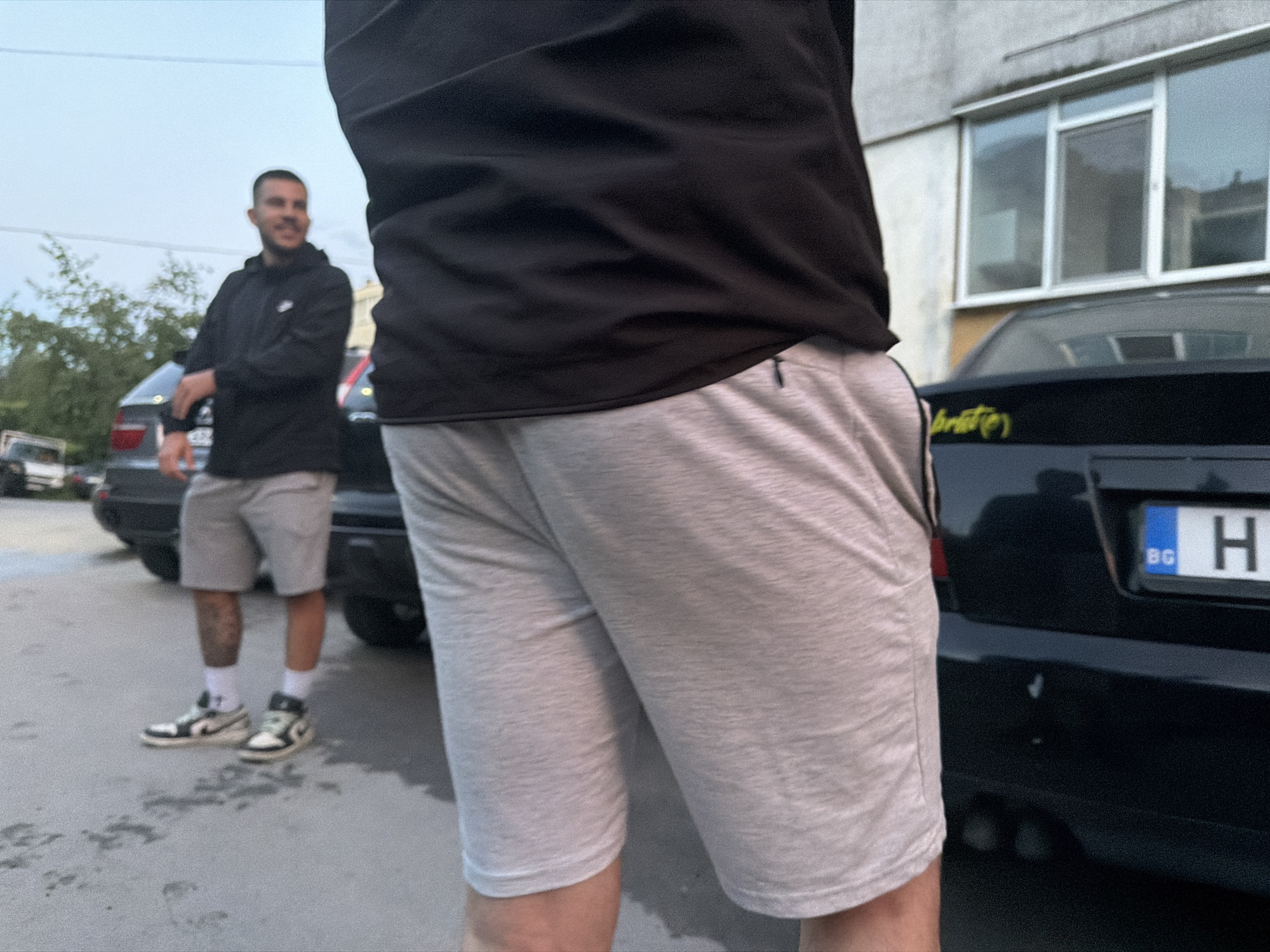 The image presents a low-angle view of a casual outdoor scene in Shumen, Bulgaria, featuring two men amidst parked cars and a building, suggesting a relaxed social interaction.
In the immediate foreground, the back of a person is visible, dressed in a black long-sleeved top and light grey athletic shorts with a small black drawstring loop on the left side.
To the left and slightly behind this foreground figure, a young man with short dark hair and a beard is seen smiling. He is wearing a black hoodie with a white Nike swoosh logo on the left chest, light grey athletic shorts, white socks, and black and white high-top sneakers. Tattoos are visible on his left calf. He leans casually against the rear of a dark-colored SUV.
The setting includes several cars; a dark car on the right displays a Bulgarian license plate, partially obscured but showing "BG H". Yellow, stylized text resembling "Sprite" is visible on its trunk lid. A light-colored, somewhat weathered building with large windows is in the background on the right. The ground is paved with asphalt or concrete, showing some damp patches.
The soft, diffused lighting suggests it is either an overcast day or late afternoon/early evening. The visible smile of the man in the background indicates a friendly or relaxed atmosphere. The overall scene captures a candid moment of two individuals conversing or casually hanging out.