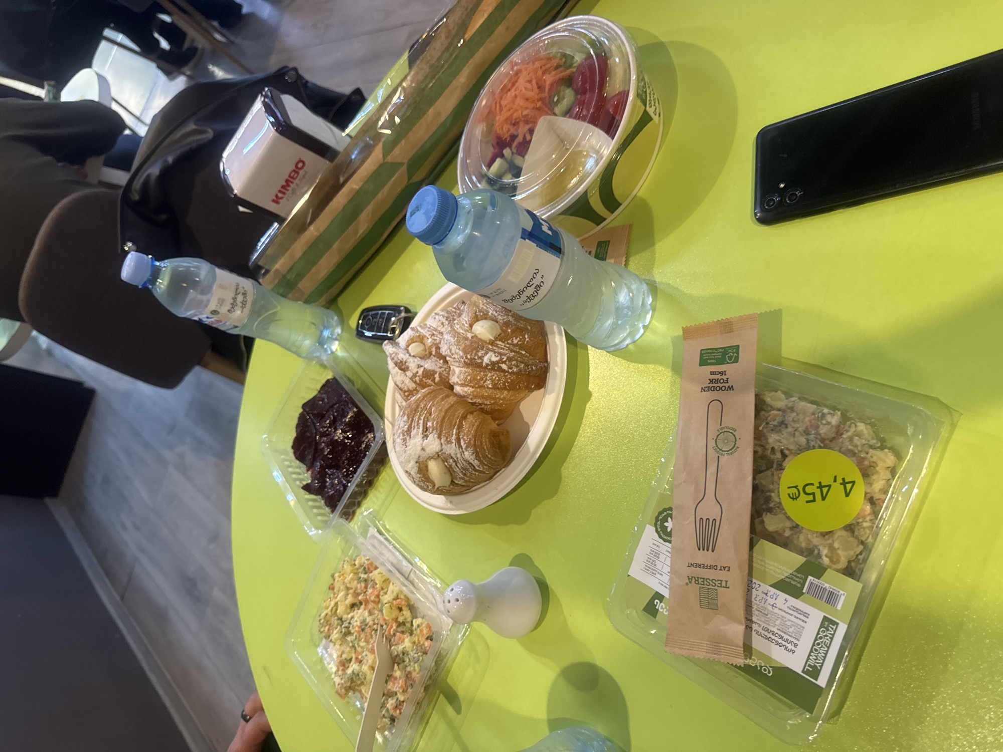 On a light green surface, likely a tabletop, there are several food items. A clear plastic container holds a salad with carrots and red vegetables. Nearby is a bottle of water with a blue cap. A white plate holds pastries, dusted with powdered sugar. There's also a clear plastic container with a dark-colored spread and another with a salad of mixed vegetables. Next to the mixed vegetable salad is a small white container. A brown paper bag, seemingly containing food, is partially visible. A black smartphone rests on the surface, next to a wrapped item and a price tag indicating 4.45. The food and packaging have labels in both English and Georgian. The brand "Kimbo" is also visible on the item and the word "TESSERA" and the price tag indicating 4.45 on the food, further indicating the location might be Tbilisi, Georgia. The presence of a brown coat and the corner of a chair suggests the setting is an indoor location, possibly a cafe or restaurant.