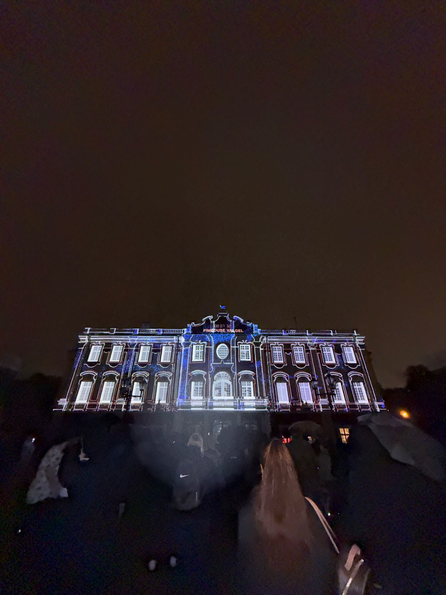 The image captures a nighttime view of a grand building illuminated by a light projection. The building, an architectural style with multiple windows and decorative elements, is bathed in blue and white light, outlining its structure and windows. On the facade, illuminated text is visible, reading "PIMEDUSE VALGEL" and "00:07:32". The scene appears to be outdoors, with dark, overcast skies above. In the foreground, the silhouettes of several people are visible, some appearing to be standing and observing the light display. The ground in the foreground is dark and appears to be a paved or grassy area. The overall impression is of a public event or exhibition taking place at night, likely in a historical or significant urban setting. The text "PIMEDUSE VALGEL" translates from Estonian to "In the Light of Darkness". While the image itself does not explicitly show Tallinn, the Estonian text strongly suggests the location.