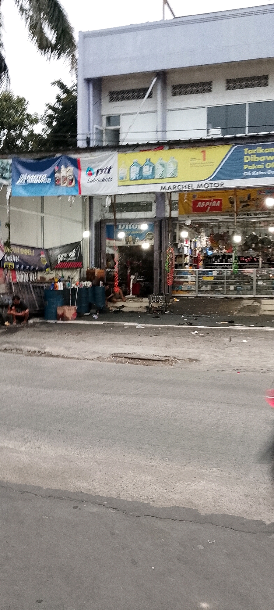 The image depicts a street-level view of a commercial building in Depok, Indonesia, likely a motor vehicle parts and service shop. The building's facade features several signs. A prominent banner above the shop entrance reads "MARCHEL MOTOR" in large letters, with a smaller sign above it showing images of oil bottles and the text "Tarikan Dibawa Pakai Oli" which translates to "Pulling Carried Use Oil". Another banner on the left side shows "MOTO SERVICE" and "ptt Lubricants". The shop entrance appears to be a doorway with a visible interior displaying shelves stocked with various products. To the left of the entrance, there are several large blue barrels and a red barrel on the sidewalk, along with a person crouching down. The street in front of the building is paved and shows signs of wear, including a pothole. The environment suggests a typical urban setting in Indonesia.