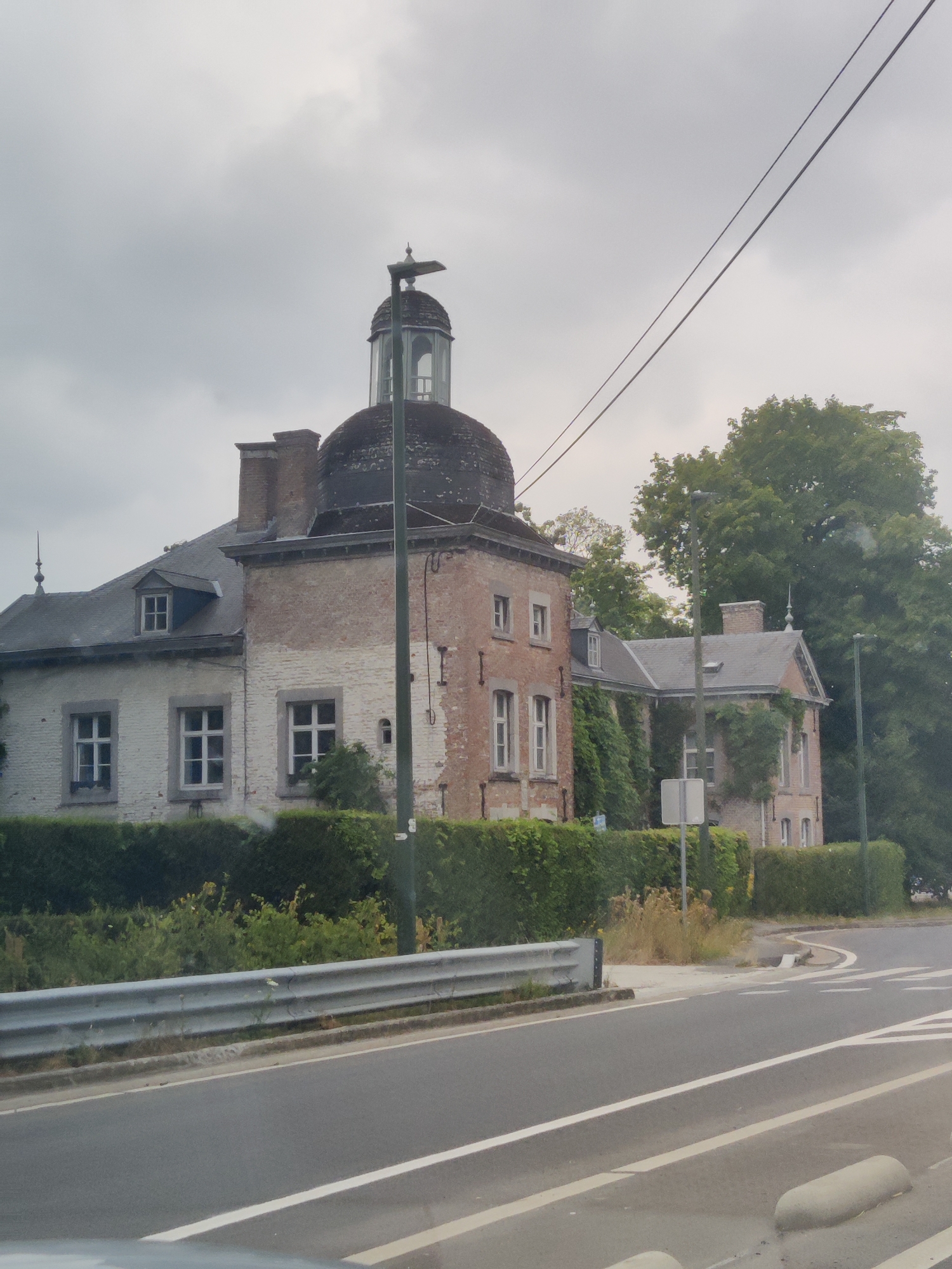The image depicts a view of the exterior of the Château de Froidcourt, located in the municipality of Nandrin, Belgium. The château is a historic building with distinct architectural features, including a prominent dome and a mix of white and red brick facades. The building is partially obscured by lush green hedges and tall trees. A road runs in front of the château, with a metal guardrail separating it from the property. Several streetlights are visible, and overhead power lines crisscross the sky, which is overcast. The scene appears to be captured from a moving vehicle, as suggested by the slight blur and the perspective of the road. There are no people visible in the image.