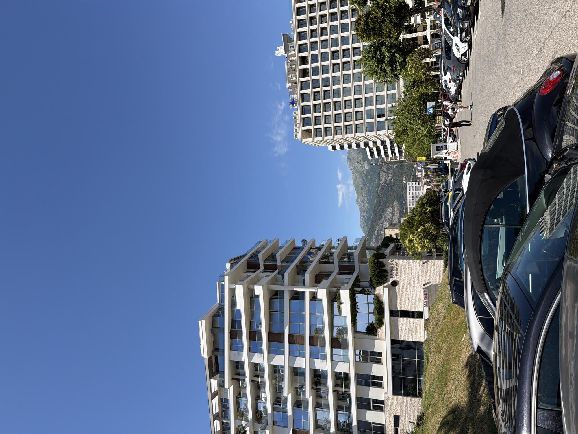 The image shows a street scene in Budva, Montenegro, on a sunny day. Two modern buildings dominate the view. The building on the left is a white, multi-story apartment complex with balconies and large windows. The building on the right is a larger, rectangular building with many windows, appearing to be a hotel or commercial establishment. In the foreground, several cars are parked on a street. Two people are walking in the distance between the buildings. The sky is clear blue, and mountains are visible in the background. The scene appears to be a resort area, given the architecture and the presence of parked cars.