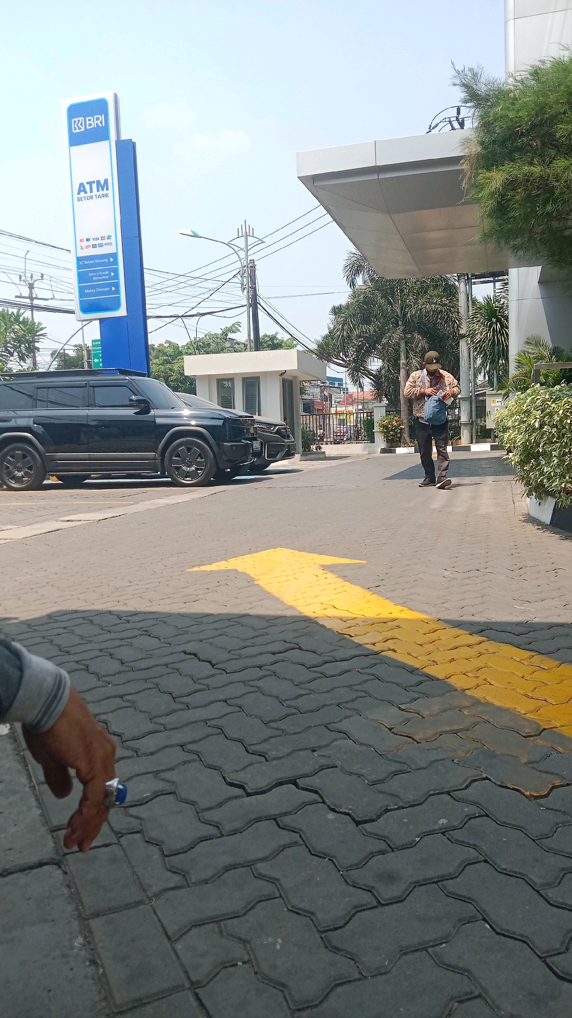 The image depicts an outdoor scene near a BRI (Bank Rakyat Indonesia) ATM and branch in Bekasi, Indonesia. In the foreground, a person's hand wearing a ring is visible, resting on a grey interlocking paver pathway. A yellow directional arrow is painted on the pathway, pointing to the right.
In the mid-ground, a black SUV is parked. Behind the SUV, a person is walking, carrying a blue bag and wearing a cap and a patterned jacket. A small white booth-like structure is also visible behind the SUV.
The background shows the BRI ATM signage, a modern building with a white overhang, trees, and utility poles with electrical wires against a bright sky. The environment suggests a commercial or financial district area within the city of Bekasi.