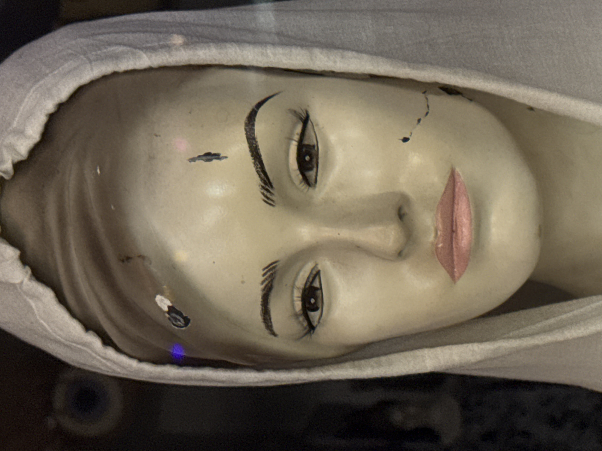 The media file displays a close-up, low-angle, tilted shot of a mannequin's face, adorned with a white cloth covering its head. The mannequin's skin tone is pale, with painted features including dark, arched eyebrows, closed eyes with painted eyelashes, and pink lips. There are several small marks and imperfections on the mannequin's face and head, suggesting age or wear. A dark, blurry background with some out-of-focus circular shapes and a faint blue light is visible. The setting appears to be indoors, possibly a display or storage area, given the mannequin and the dark, indistinct surroundings. The lighting is uneven, casting some shadows on the mannequin's face. The image does not show any specific activity or event, focusing solely on the static appearance of the mannequin. There is no visible text in the media file. The location context provided is Karon, Thailand, but there are no visual cues within the image to confirm this.