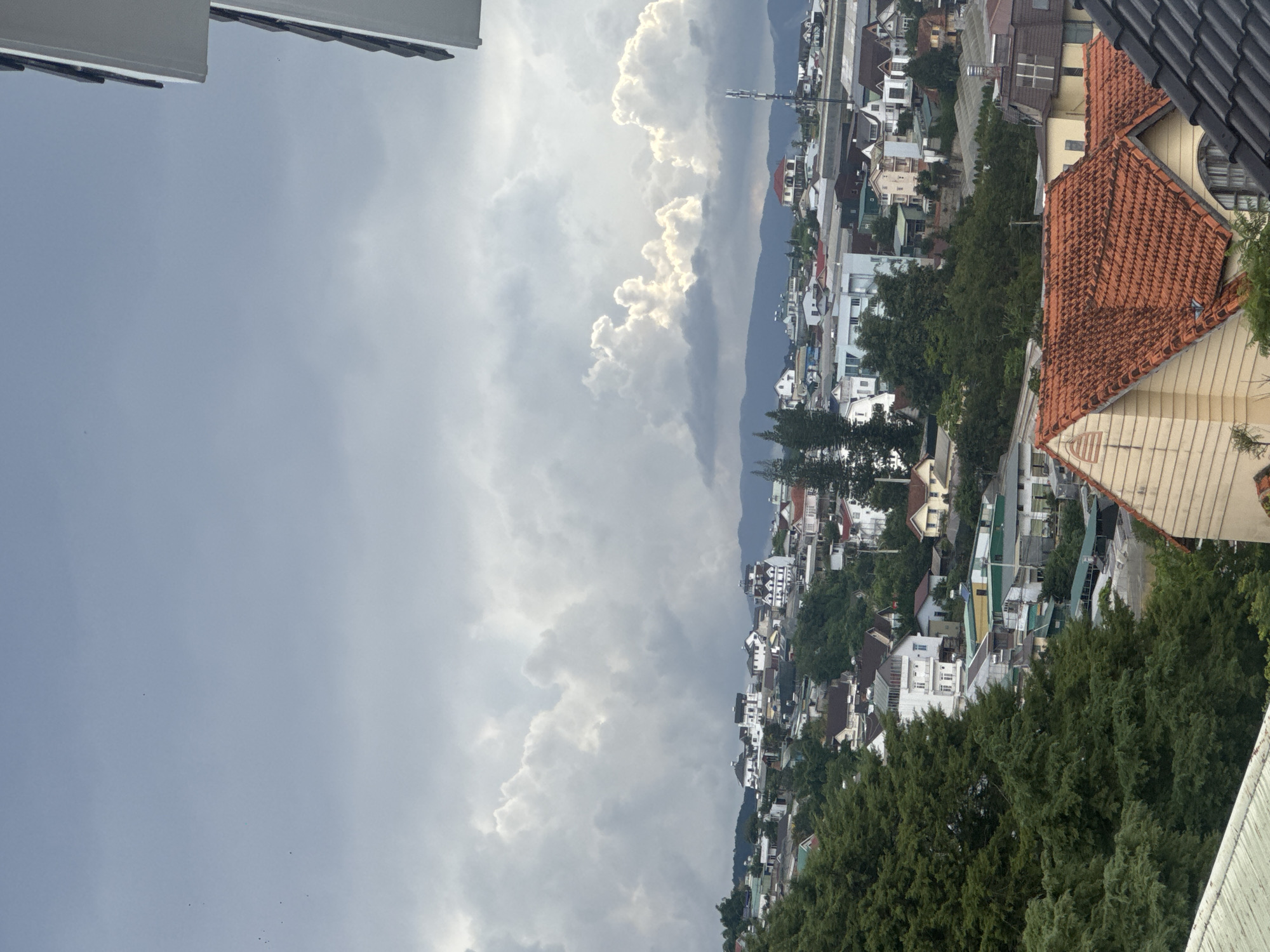 The image depicts an aerial view of a hillside city, likely Lam Vien - Da Lat, Vietnam, under a dramatic, cloudy sky. The foreground is dominated by the angled rooftops of several houses, one with a bright orange-tiled roof and a pale yellow-sided structure. To the left, a dense canopy of dark green pine trees covers a significant portion of the frame, obscuring the lower hillside.
Beyond the immediate foreground, the landscape transitions into a densely packed urban environment of houses and buildings, varying in architectural style and color. Many structures feature white or light-colored facades, interspersed with darker roofs and occasional splashes of red. The buildings are nestled amongst lush green trees and vegetation, indicating a city integrated with nature.
In the middle ground, the terrain rises, revealing more buildings and streets winding through the hillside. The background showcases a range of mountains under the expansive, overcast sky. The clouds are a prominent feature, with some areas illuminated by sunlight breaking through, creating a contrast between bright white formations and darker, more ominous clouds. A cell tower is visible in the distance, suggesting modern infrastructure within the city. The overall impression is of a cool, possibly humid climate, common in Da Lat's mountainous region. No people are visible in the image.