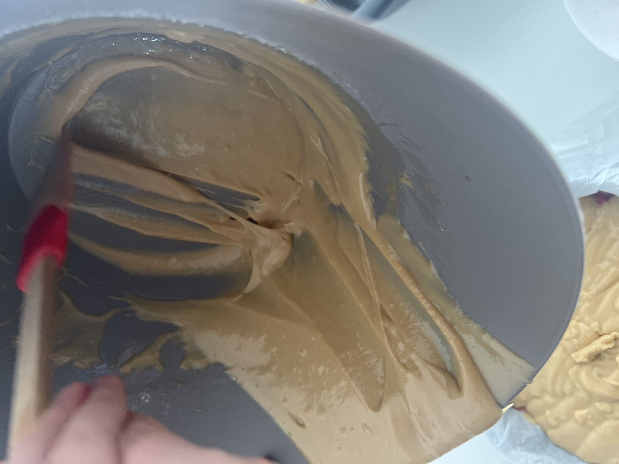 A close-up, high-angle shot depicts a gray mixing bowl nearly full of a light brown, viscous substance, possibly batter or frosting. A spatula with a red tip and wooden handle is partially visible on the left, held by a blurred hand. The substance clings to the sides of the bowl in streaks and pools in the bottom. A small section of similar substance is visible on a white surface to the right, likely being poured or scraped out. The lighting is bright and even. There is no indication of the image being taken in Mogán, Spain.