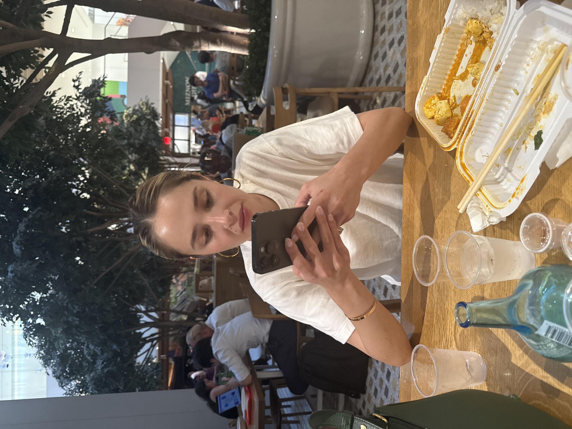 A young woman with blonde hair pulled back is seated at a wooden table, looking down at her smartphone which she holds with both hands. She wears a white t-shirt, large hoop earrings, and a thin bracelet on her left wrist. In front of her are three empty plastic cups and a partially consumed meal in a takeout container, with chopsticks resting on it. A green handbag is partially visible to her left, and a glass bottle with a blue tint is in the foreground. Behind her, other patrons are visible at tables, some with laptops. The scene appears to be indoors, possibly a food court or dining area within a large building, with tall trees and potted plants present. A sign in the background reads "MIZNON". The overall lighting suggests an indoor, possibly well-lit environment.