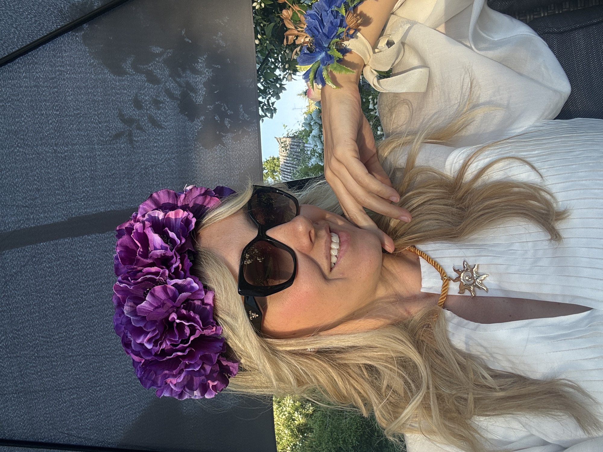 The image shows a woman lying down outdoors. She has blonde hair, is wearing sunglasses, a white top, and a flower crown made of purple flowers. She also has a flower bracelet on her wrist. The woman is smiling and appears relaxed. In the background, there are trees and a building. The lighting suggests it is daytime, with sunlight filtering through the trees. The overall scene conveys a sense of leisure and enjoyment. The location is Carnikava, Latvia. There is visible text on the sunglasses frame, which appears to be "PRADA".