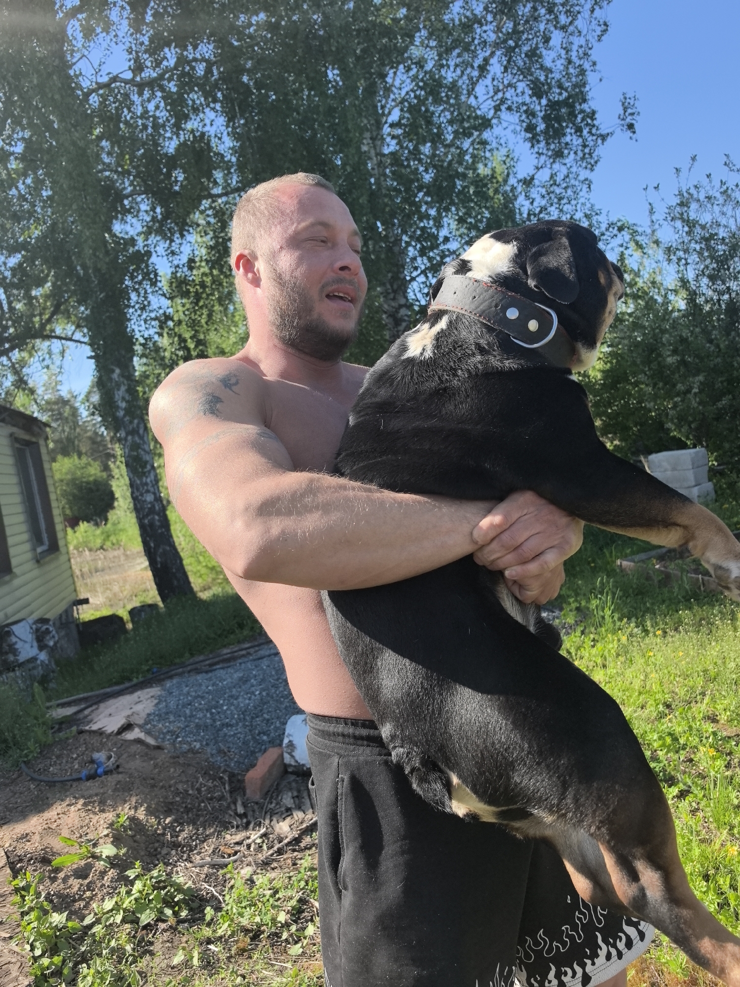 A shirtless man with short hair and a beard is holding a large black and brown dog in his arms outdoors. The dog has a thick black collar with silver accents. The man is wearing black athletic shorts with a white flame pattern along the hem. A weathered, light yellow building is visible in the background on the left. Trees and green foliage fill the background. A dirt and gravel area is visible in the foreground. The location appears to be an outdoor yard or garden. The city is not clearly visible in the image.