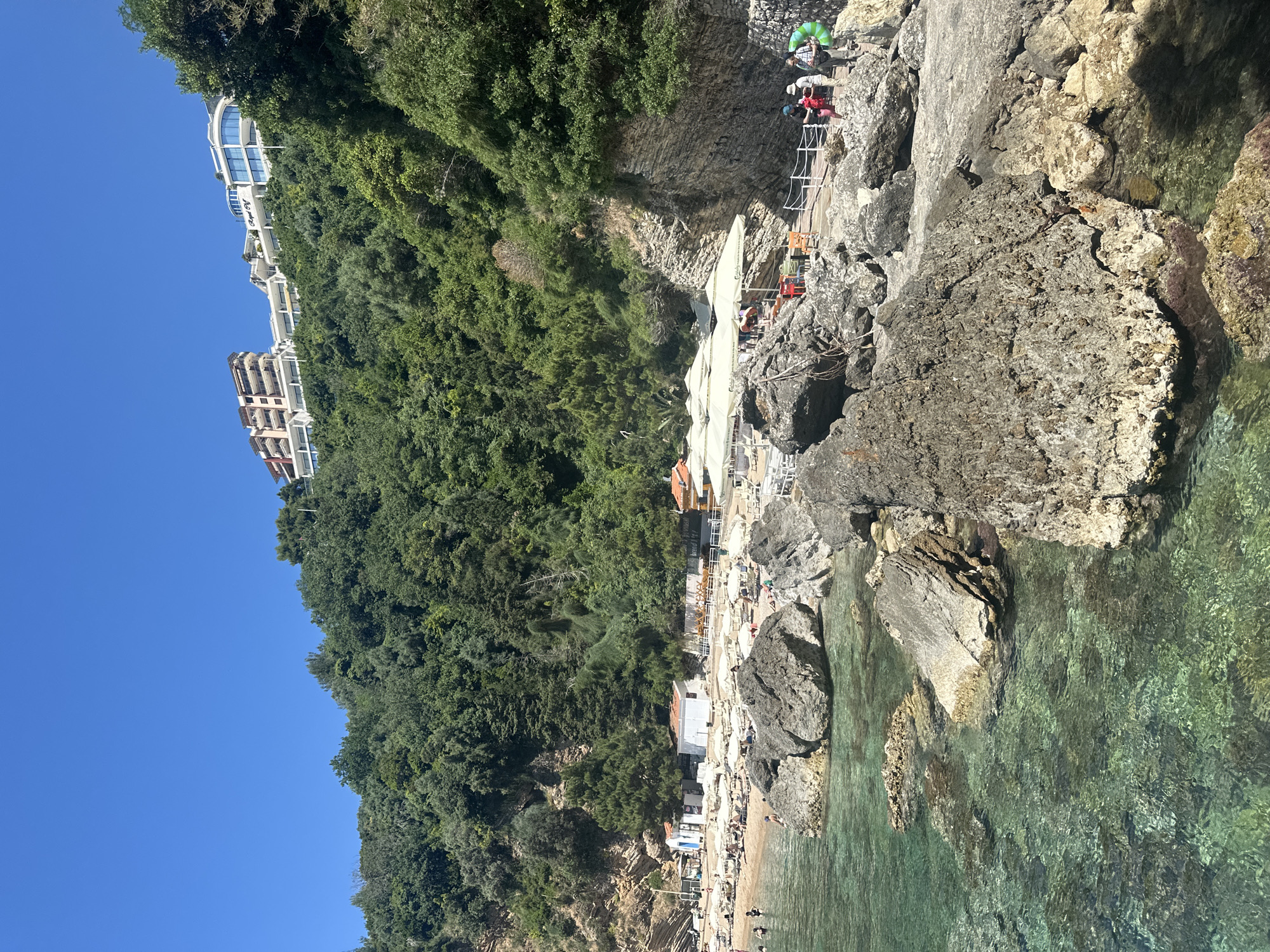 The image shows a beach scene near Budva, Montenegro. The foreground features a rocky coastline with clear turquoise water. Further back is a sandy beach populated with people, sun umbrellas, and lounge chairs. Above the beach, a steep, verdant hillside rises, covered in lush trees and foliage. On the upper part of the hill, there are several multi-story buildings with white facades, seemingly hotels or apartments. A narrow pedestrian walkway is visible connecting different points along the rocks, with people walking and standing on it.