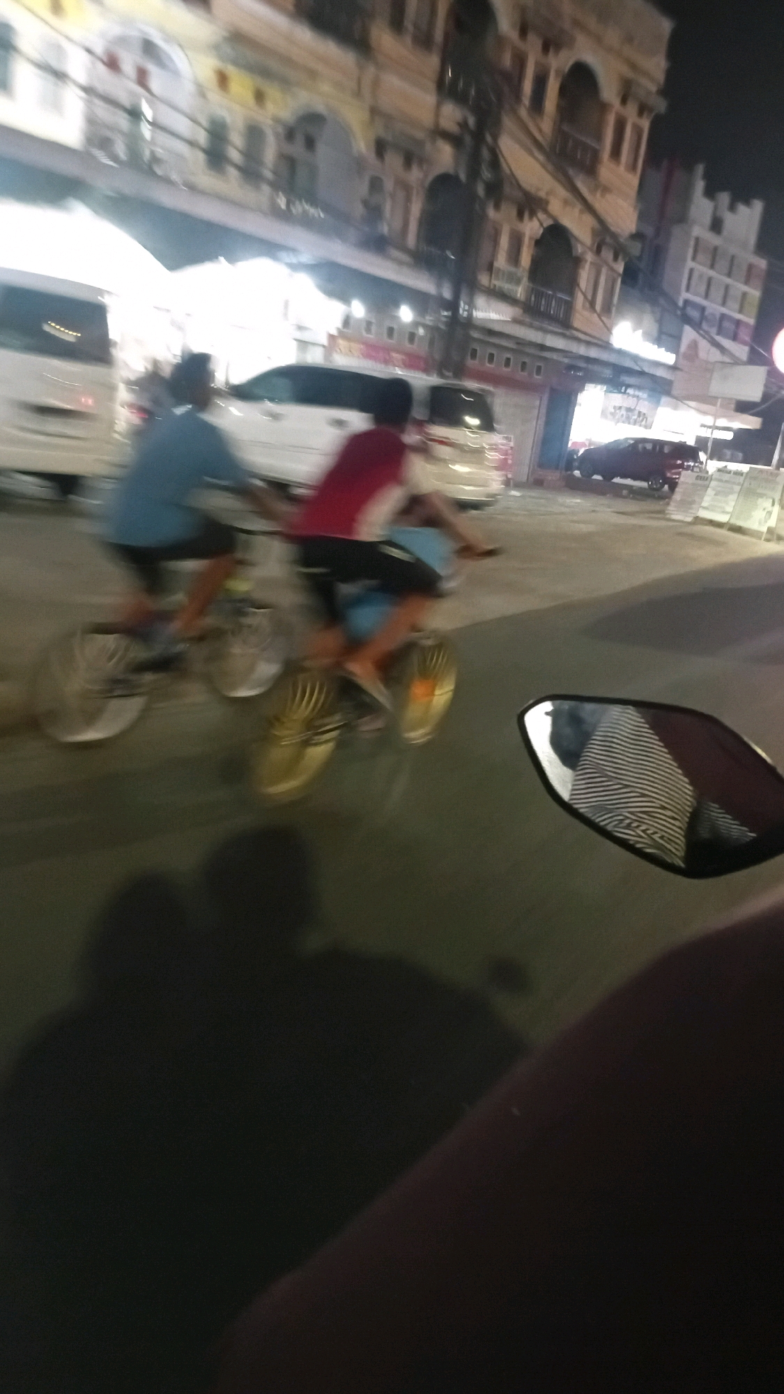 The image depicts a night scene on a city street in Jambi, Indonesia, likely in an urban commercial area. Two individuals are riding bicycles in motion, causing blur. The person in the foreground is wearing a red shirt and dark shorts, while the person behind them wears a blue shirt and dark shorts. They are riding on a paved road with a faint shadow cast on the asphalt. In the background, there are buildings with architectural details, suggesting older structures, possibly commercial establishments or residential units above shops. Several vehicles, including a white van and a dark-colored SUV, are parked or driving in the background. A motorcycle's rearview mirror, reflecting a striped pattern, is prominently visible in the lower right corner of the frame, indicating the photographer's perspective from a moving motorcycle. The lighting suggests the presence of streetlights and illumination from the buildings. The overall impression is a busy, albeit somewhat blurry, snapshot of nighttime urban activity.