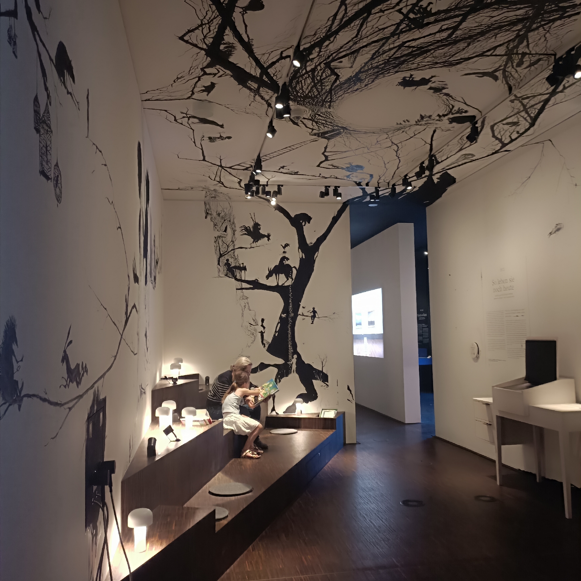 The image depicts an exhibition space, likely within a museum or gallery, featuring striking black and white murals on the walls and ceiling. The murals appear to be intricate drawings of trees and various figures, giving the space a surreal or fantastical atmosphere. In the foreground, a woman and a young girl are seated on a tiered, wooden bench, engaged in reading a book. The girl is pointing at the book, and the woman is looking at it with her. Several small, modern lamps are strategically placed on the bench and nearby, providing illumination.
Further into the space, there is a projection screen displaying an image of buildings. To the right, a white desk-like structure with drawers and a compartment is visible, along with some printed text on the wall above it. The floor is made of dark wood. The lighting is atmospheric, with spotlights directed at the ceiling and artwork.
This scene is located at the Jüdisches Museum Frankfurt (Jewish Museum Frankfurt), specifically in their historical exhibition "Befreit und verwehrt. Frankfurt und die Juden seit 1945" which opened in 2022. The artwork on the walls is by the artist Hélène M. V. C. M. P. D. and relates to the exhibition's themes. The specific area shown in the image is the children's and family area within the exhibition.
