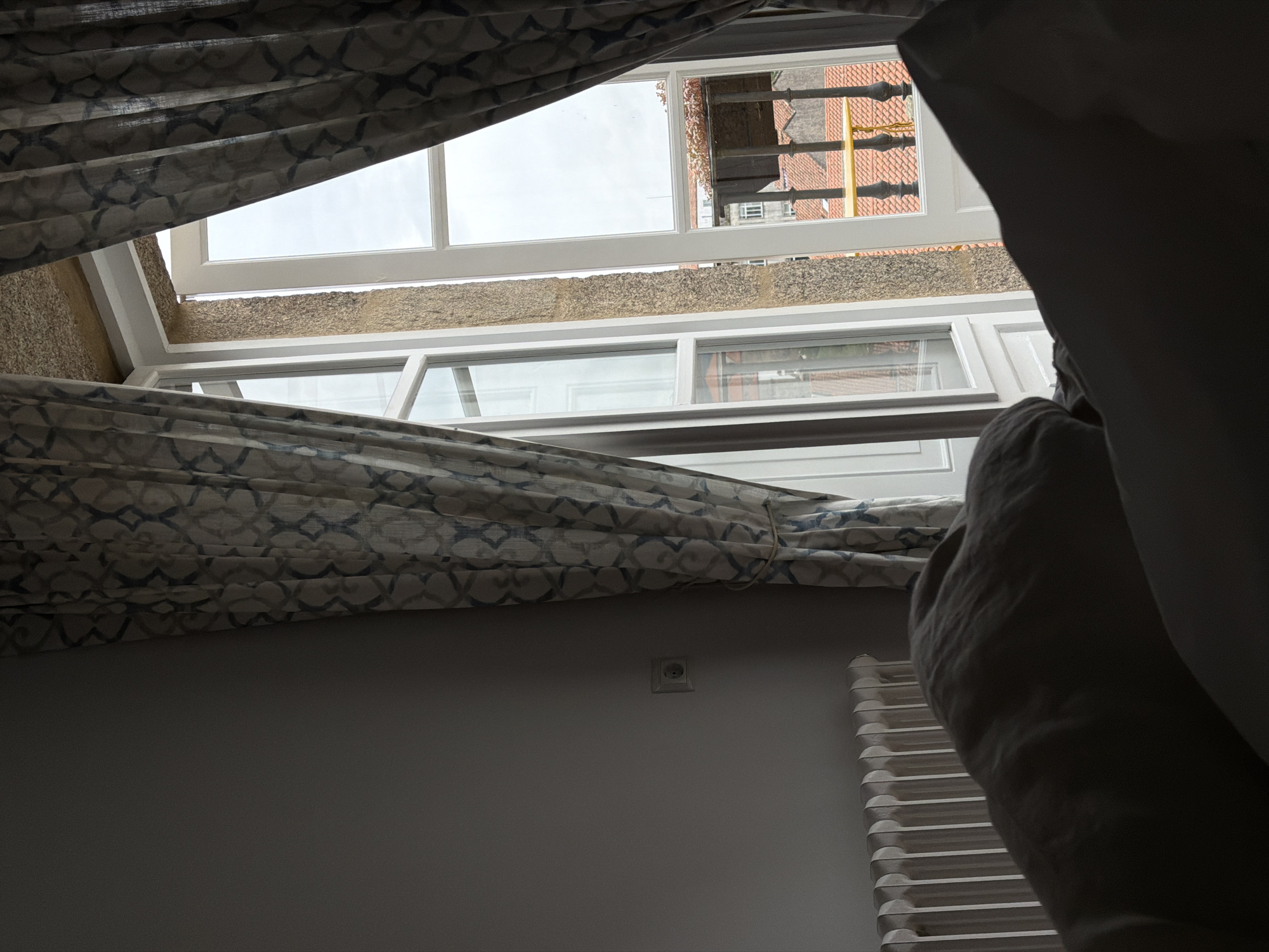 The image shows a view from inside a room in Santiago De Compostela, Spain, looking out through an open window. The window has a white frame and is divided into panes. Outside, scaffolding is visible on a red-tiled roof. A patterned curtain partially covers the window, with folds and creases indicating movement or being drawn back. In the lower part of the frame, a section of a radiator is visible against a plain, light-colored wall, with a small electrical outlet positioned above it. A large, out-of-focus object, likely part of bedding or clothing, is present in the right foreground, obscuring part of the view.
