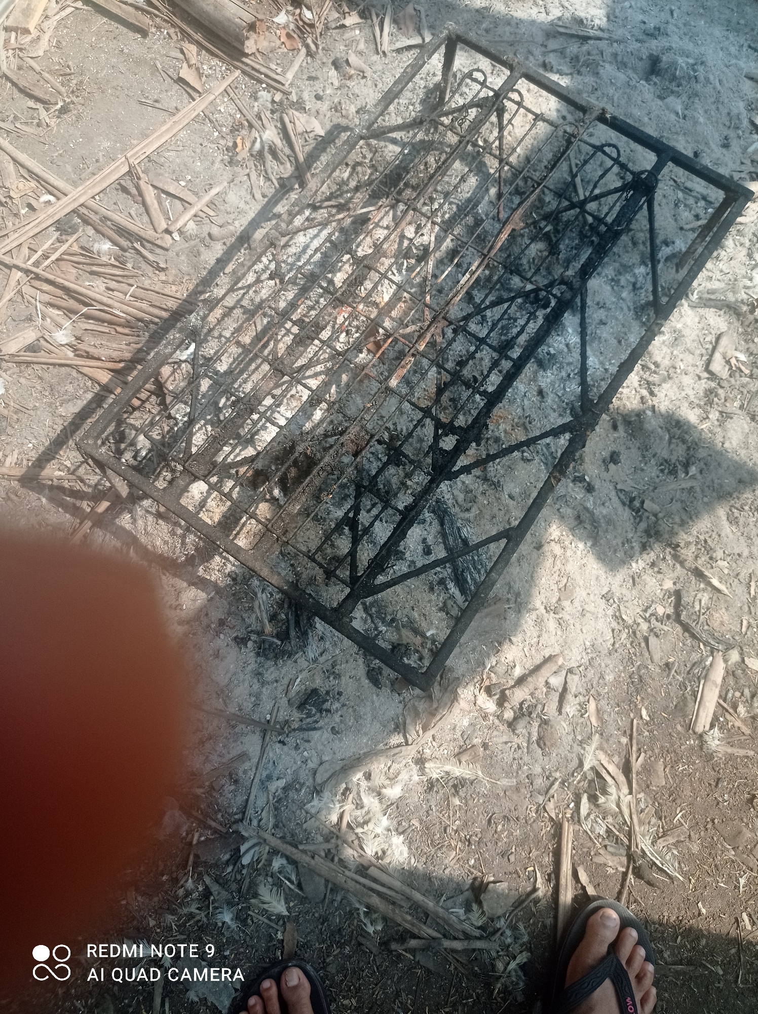 The image depicts an outdoor scene on the ground, likely in Cipawon, Indonesia, characterized by dry earth, scattered debris, and a prominent metal grill. The ground is covered in a mix of sand, dirt, and what appears to be ash and burnt material. Numerous dry, elongated organic elements, possibly palm fronds or similar plant matter, are strewn across the surface, along with some white feathers.

In the center of the frame, a rectangular metal grill lies horizontally. It is constructed with a black metal frame and a grid of thinner metal bars. The grill shows signs of heavy use and scorching, with a significant amount of soot and residue adhering to its structure. The grill appears to be empty, suggesting it was recently used for cooking or burning.

The lighting suggests a sunny day, casting distinct shadows across the scene, including the grill and some of the organic debris. In the lower left corner, a large, out-of-focus reddish-orange object obscures part of the scene. In the lower right corner, a person's bare foot wearing a black flip-flop is visible, standing on the ground, indicating human presence. The lower portion of the image also shows a watermark with "REDMI NOTE 9 AI QUAD CAMERA." The overall impression is one of a rustic, possibly recently cleaned-up area, with a focus on the burnt grill as a central element.