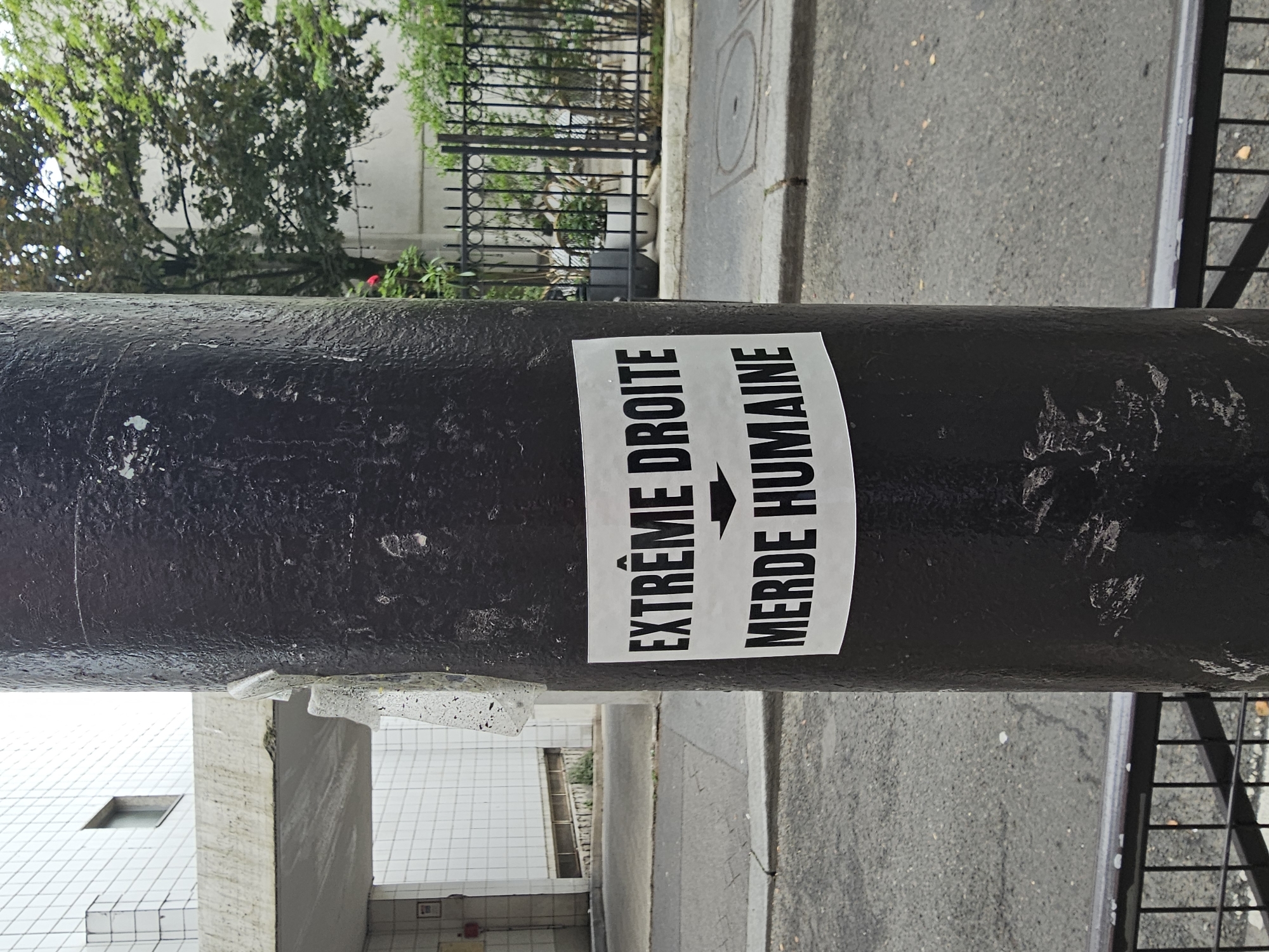 A black bollard, situated on a Paris street, displays a sticker bearing the French phrase "EXTRÊME DROITE" followed by an arrow pointing to the phrase "MERDE HUMAINE." The surrounding environment includes trees, a fence, an adjacent building with tilework, and a paved street.