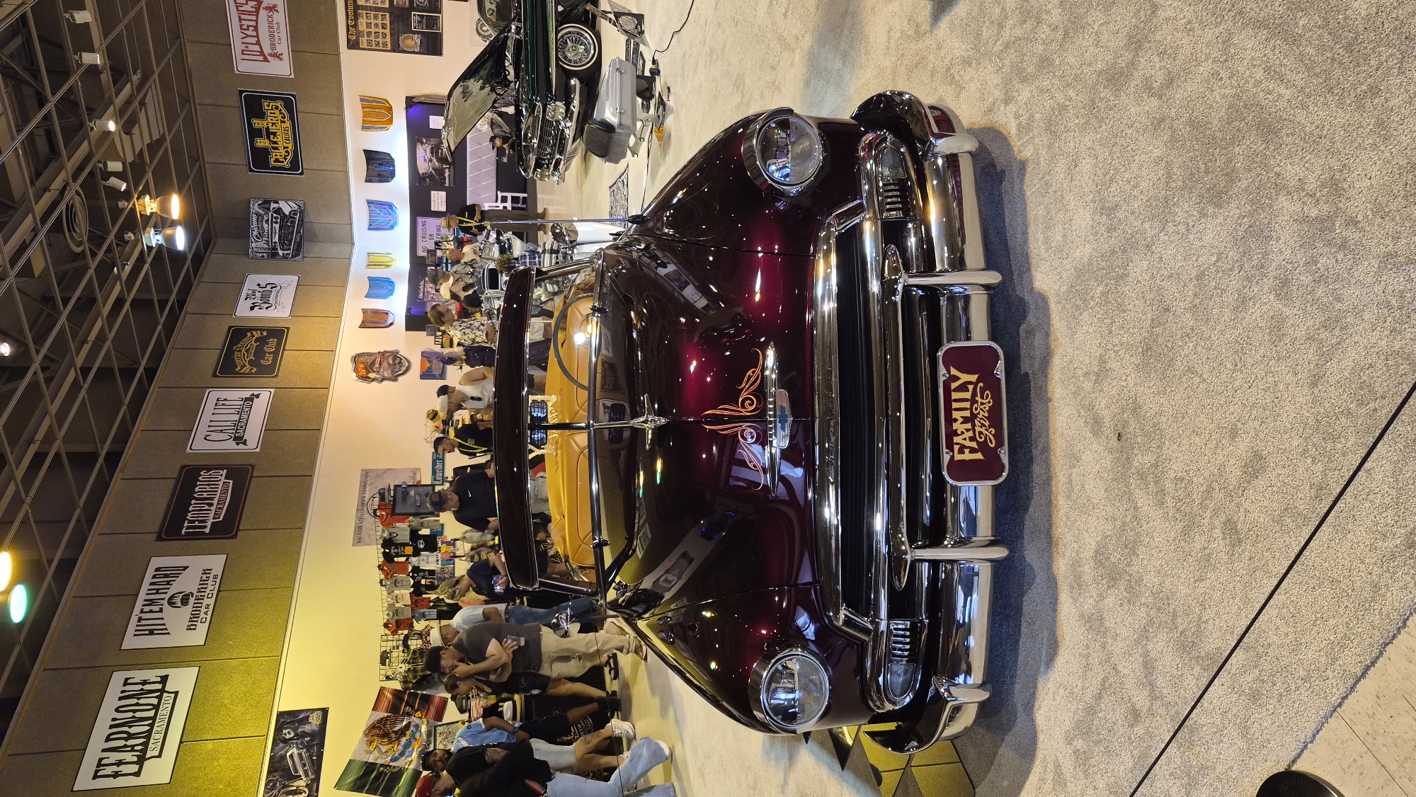 The image shows an indoor event, likely a car show, with several classic cars on display and people browsing. The setting appears to be a convention center or exhibition hall, indicated by the high ceilings, lighting fixtures, and the overall layout. The prominent vehicle in the foreground is a dark red convertible with a tan interior, featuring a "FAMILY FIRST" license plate. In the background, another car, a dark green sedan with its hood open, is visible. A wall behind the cars is adorned with various signs and banners, some of which mention "Sacramento" and "Car Club," suggesting a connection to the car culture within the city. The floor is covered with carpeting. People are dressed in casual attire and are standing, walking, and observing the vehicles. The lighting is bright, illuminating the exhibition space.