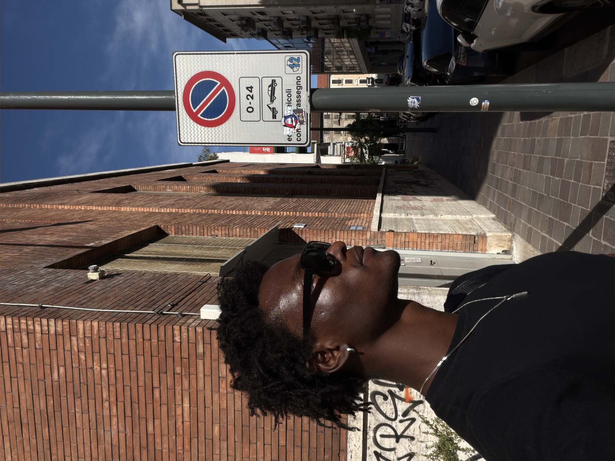 An outdoor, medium shot shows a Black man in profile, facing left, wearing sunglasses, a black t-shirt, and a silver necklace. He stands on a sidewalk paved with brick pavers, next to a brick building. A lamppost with a parking sign stands in front of the man. The sign indicates a no parking zone from 0-24 hours and includes symbols of a tow truck and a car with a ticket. The background shows a street with parked cars, more buildings, and a blue sky. The man's hair is dark and curly, and he has an earring in his left ear. Graffiti is visible on the wall behind him. The overall scene appears to be in a city, likely Milan, Italy, given the style of the building and the parking sign.