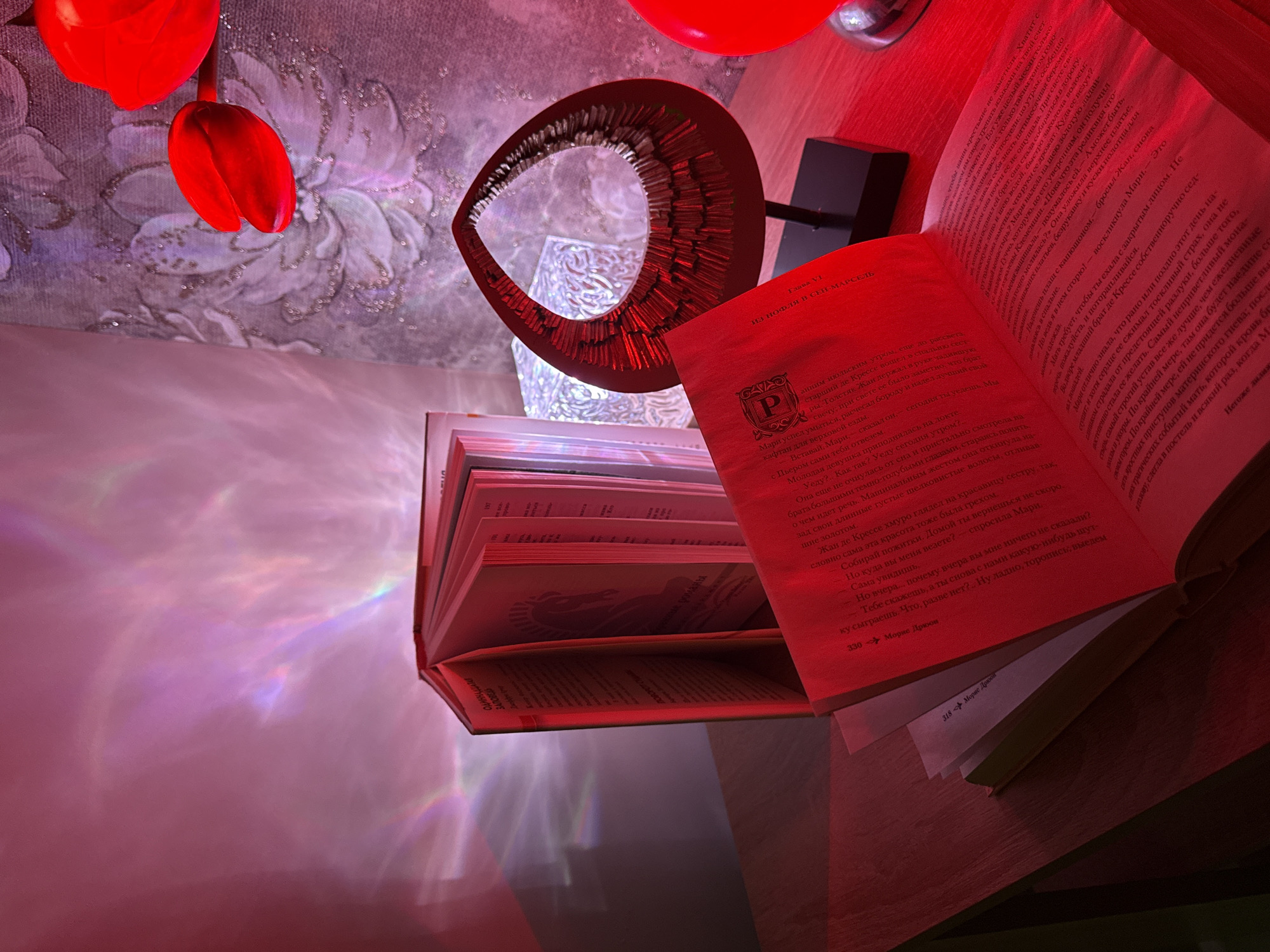 An indoor scene is depicted with a strong red and pink ambient light, illuminating books, decorative items, and a textured background. In the foreground, an open book with Cyrillic text rests atop a stack of other books, partially revealing its pages and spine. The red light casts a warm glow on the pages, making some parts appear darker red and others brighter.
Above the books, a distinctive metallic, ring-shaped object with a textured, possibly spiky, inner surface catches the light, glowing intensely red. Behind this object, a small, square, black item is visible, possibly a base for the metallic sculpture or another device. To the left, two red, artificial tulip-like flowers hang down, vibrant against a softly illuminated background featuring a floral pattern with subtle glitter or sequins. A red balloon is also partially visible at the top.
The red lighting creates iridescent, prismatic reflections and streaks of light across the wall or surface behind the books, adding a dreamy or ethereal quality to the scene. These light patterns range from pink to hints of blue and green.
Visible text from the open book includes titles and names in Cyrillic script: "ОДИННАДЦАТАЯ ЗАПОВЕДЬ" (The Eleventh Commandment), "Глава VI" (Chapter VI), and "Морис Дрюон" (Maurice Druon), indicating the author. Page numbers "330" and "318" are also visible. A stylized letter "P" logo is printed on the page. The narrative mentions characters like "Мари" and "Жан," and references "early July morning, before dawn" and "by candlelight," although the image itself appears to be artificially lit.