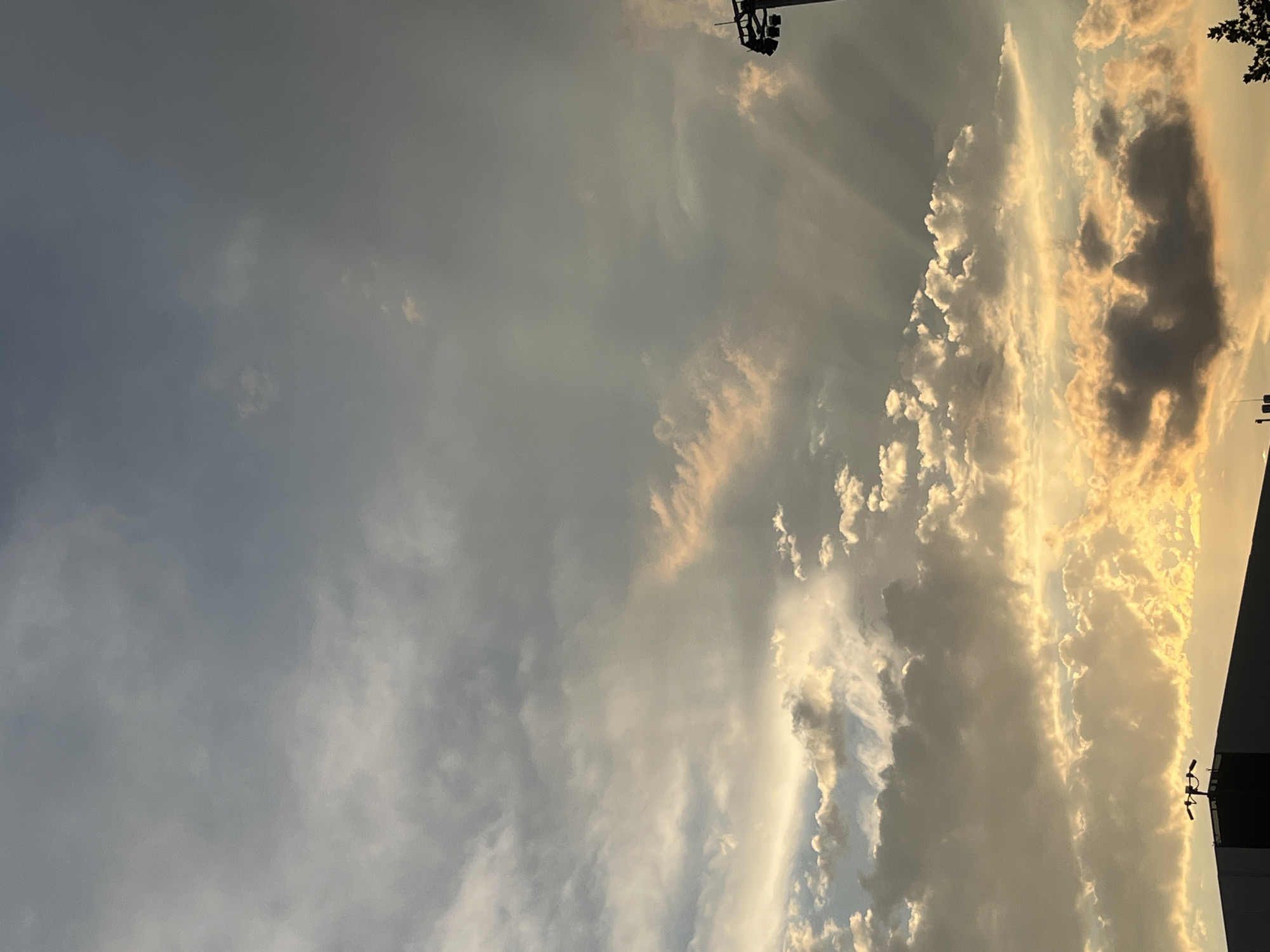 The media file depicts a sky filled with clouds, illuminated by the golden light of either sunrise or sunset. The clouds are varied in texture and color, ranging from soft white and gray to dramatic dark gray and bright yellow where the sun's rays are most intense. Some clouds appear wispy and scattered, while others are dense and voluminous, creating a dynamic interplay of light and shadow.
Notable details include visible sunbeams or crepuscular rays emanating from behind some of the clouds, indicating a low sun angle. In the upper left portion of the frame, a silhouette of what appears to be a lighting rig or fixture can be seen against the sky. In the lower right, a dark, angular structure, possibly a building or part of a structure, is visible, with a smaller silhouetted object that might be a camera or sensor on top. A few dark leaves from a tree are also visible in the upper right corner.
The overall mood is serene and awe-inspiring, emphasizing the natural beauty of the sky at a particular time of day. The lighting suggests either early morning or late evening. Given the location context of Krasnodar, Russia, the scene captures a typical atmospheric condition that could be observed in this region. No visible text is present in the image.