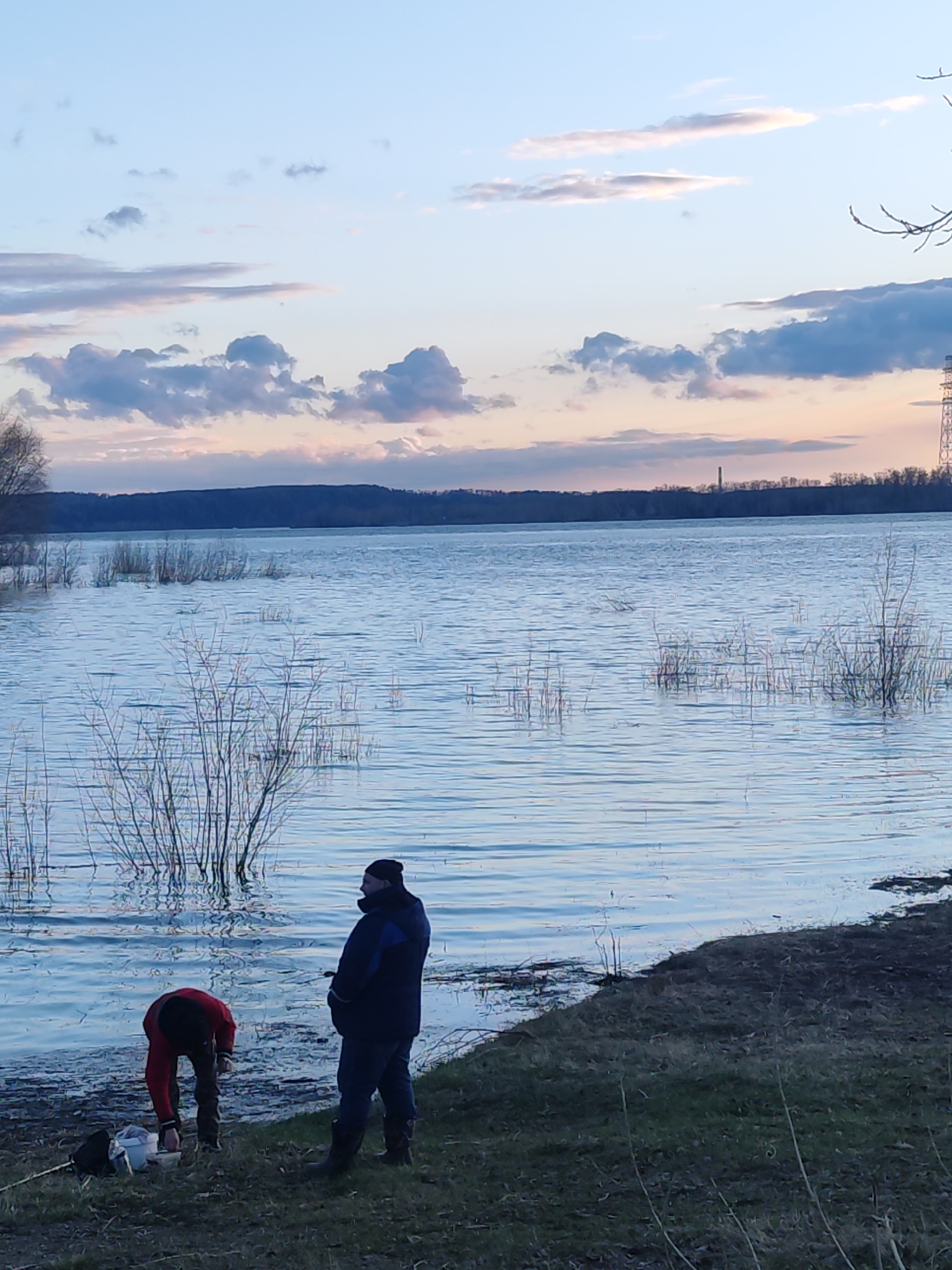 The image captures a serene outdoor scene at twilight, likely dawn or dusk, in Chaikovsky, Russia. Two individuals are positioned on the grassy bank of a large body of water, which appears to be a lake or a wide river.
In the foreground, to the right, a person in a dark blue jacket, dark trousers, and a black beanie stands upright, facing the water. To their left, closer to the water's edge, another individual dressed in a bright red jacket and dark trousers is bent over, seemingly attending to items on the ground, possibly fishing gear or bait, next to a small white container and a dark bag.
The expansive water body exhibits gentle ripples and reflects the soft light from the sky. Sparse, leafless vegetation, possibly dormant bushes or reeds, emerges from the water near the shore, suggesting a late winter or early spring setting. The background reveals a distant, tree-covered shoreline stretching horizontally across the horizon. A tall, slender industrial chimney or tower is visible on this distant land, along with what appears to be a power line tower further to the right, indicating some degree of human development near the natural landscape.
The sky is painted with delicate hues of pale blue, soft pink, and muted purple, characteristic of the early or late hours of the day. Scattered grey clouds drift across the upper sky. The overall atmosphere is calm and quiet. There is no visible text in the image.