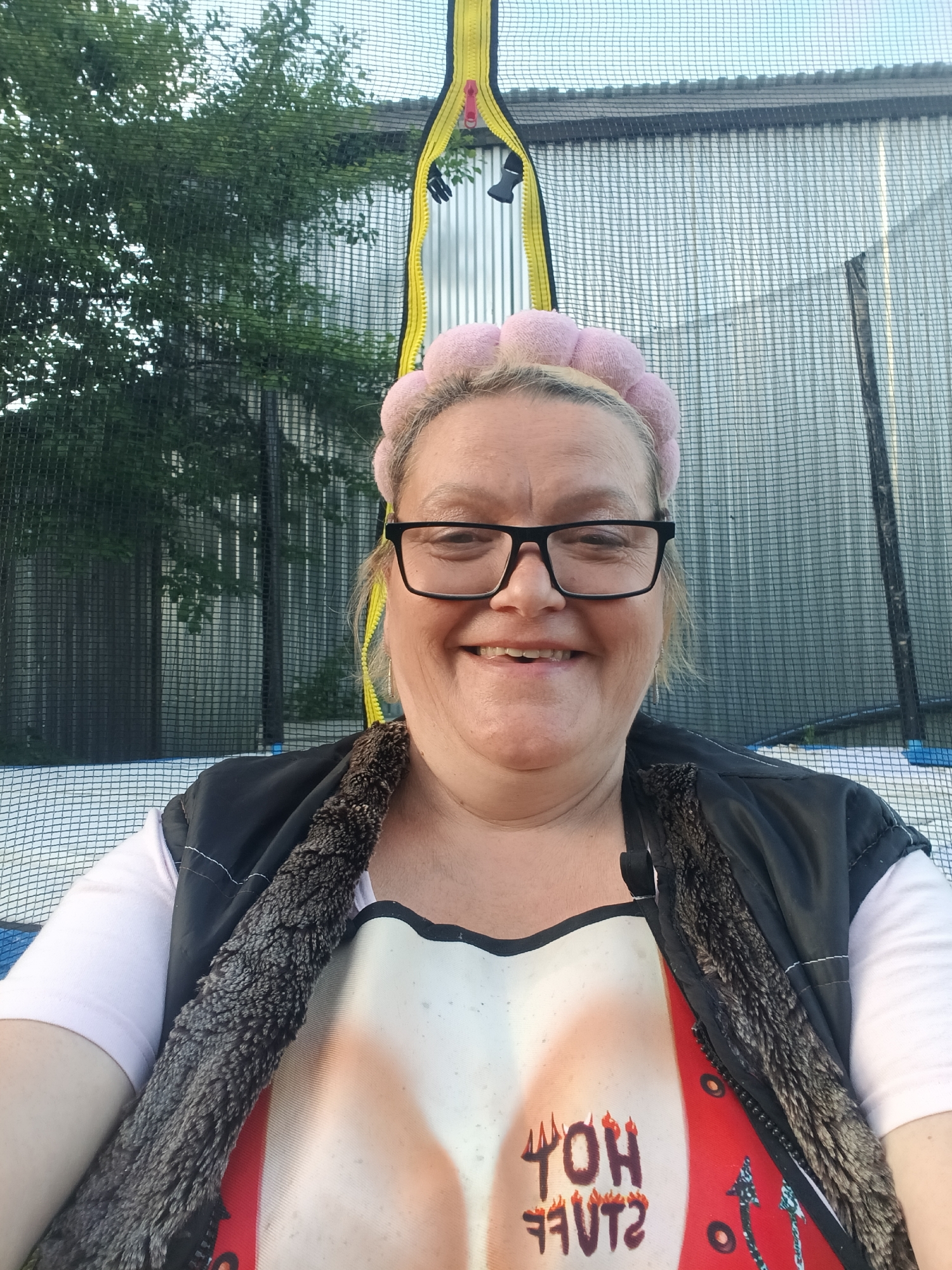 A medium close-up selfie captures a smiling woman with short blonde hair and black-rimmed glasses, wearing a pink padded headband. She is dressed in a light-colored short-sleeved top, a dark vest with a textured, fluffy brown lining, and an apron or bib over it. The apron features a humorous print of a red top with a low neckline and a white overlay, on which the text "HOT STUFF" is visible upside down in a fiery font.
The woman is positioned inside a trampoline safety enclosure, with the black mesh netting clearly visible in the foreground and background. To her left, green tree foliage is visible through the netting, while to her right, a corrugated metal fence or building can be seen. A bright yellow zippered safety strip for the trampoline enclosure runs vertically on the right side of the frame, featuring a red zipper pull. The lighting suggests it is daytime, with clear weather conditions. The woman's broad smile conveys happiness and a relaxed mood. The setting appears to be an outdoor residential or garden area, possibly in Красный Маяк, Russia.