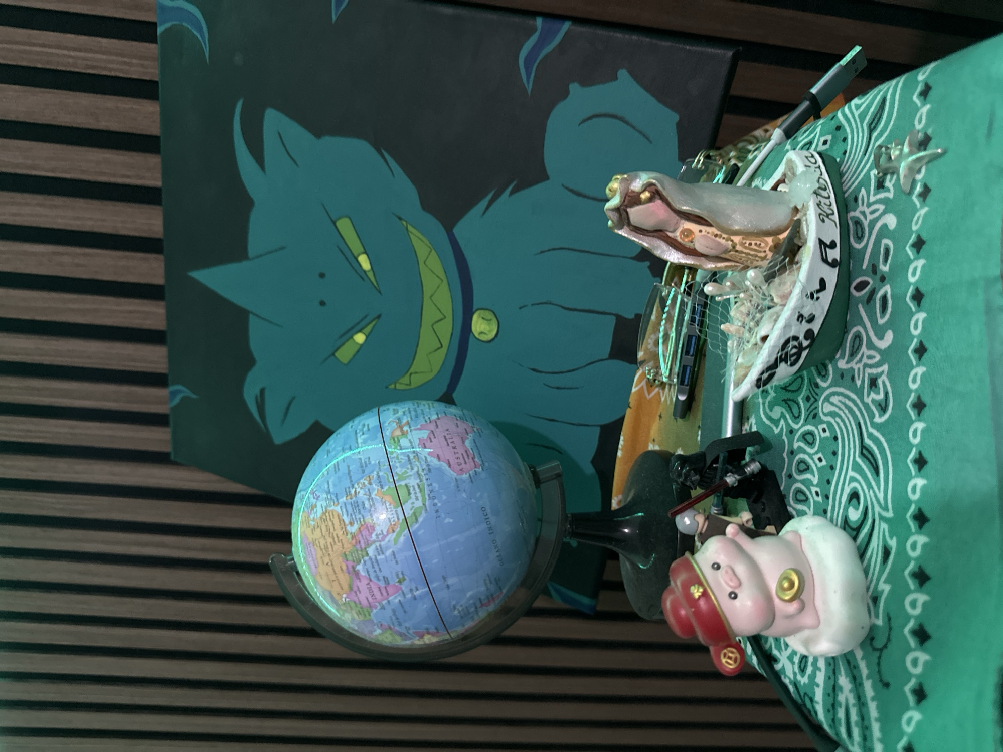 This image captures an indoor still life arrangement on a surface covered with a teal green patterned fabric, located in Sabana de Guacuco, Venezuela. In the background, a dark teal canvas painting featuring a stylized, menacing feline-like creature with sharp green eyes, a wide jagged smile, and a yellow circular collar pendant leans against vertical wooden paneling.
On the left, a small, illuminated desktop globe on a black stand displays political boundaries, including India, Malaysia, Indonesia, and Australia. On the patterned fabric, a light pink pig figurine wearing a red hat with gold accents holds a gold coin while resting on a white cloud-like base. To its right, two miniature figures resembling Lego minifigures are present: a black Darth Vader figure holding a red lightsaber, and a lighter-colored figure behind it. Further right, a white and green miniature boat figurine, with "Kitsada" partially visible on its side, contains small seashells, pebbles, and a net. Rising from the boat is a stylized, internally illuminated female figure with gold and white details. Additional small objects on the fabric include a white golf ball-like sphere, a pen, a USB stick, and a silver starfish figurine. The scene is moderately lit, highlighting specific objects with internal illumination.