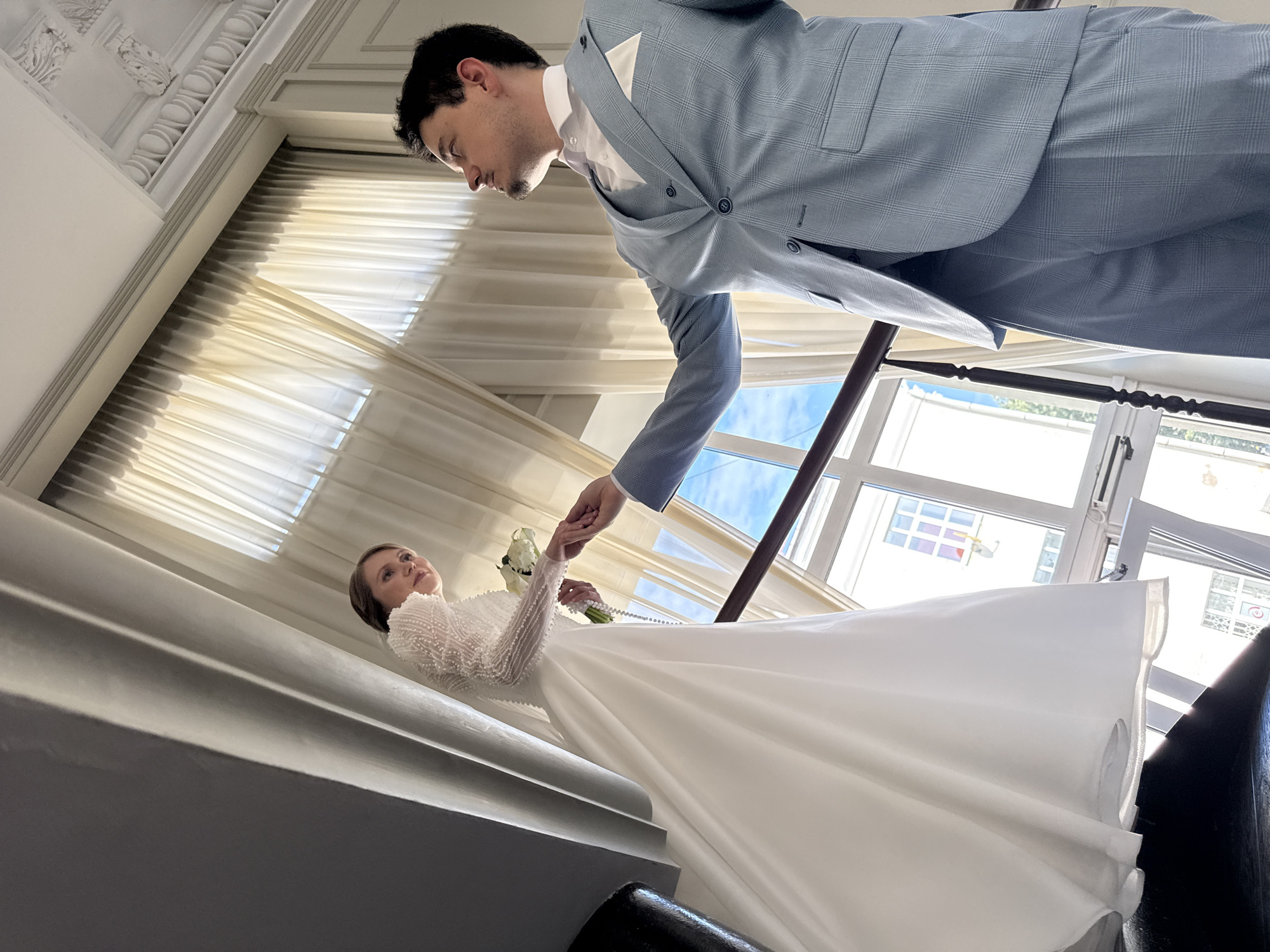 The image depicts a wedding scene on a staircase, with a bride in a white gown and a groom in a light blue suit. The bride, on a higher step, extends her left hand down to the groom. She is looking to her left, with her hair pulled back and a bouquet in her right hand. The groom, on a lower step, is reaching up to hold her hand, looking upwards towards her. He is wearing a white shirt, a vest, and a patterned suit jacket with a white pocket square. The staircase has a dark wooden railing and is illuminated by natural light streaming through large windows behind them, which offer a view of buildings and the sky. The architecture of the building features ornate ceiling moldings, suggesting an indoor setting within a city.