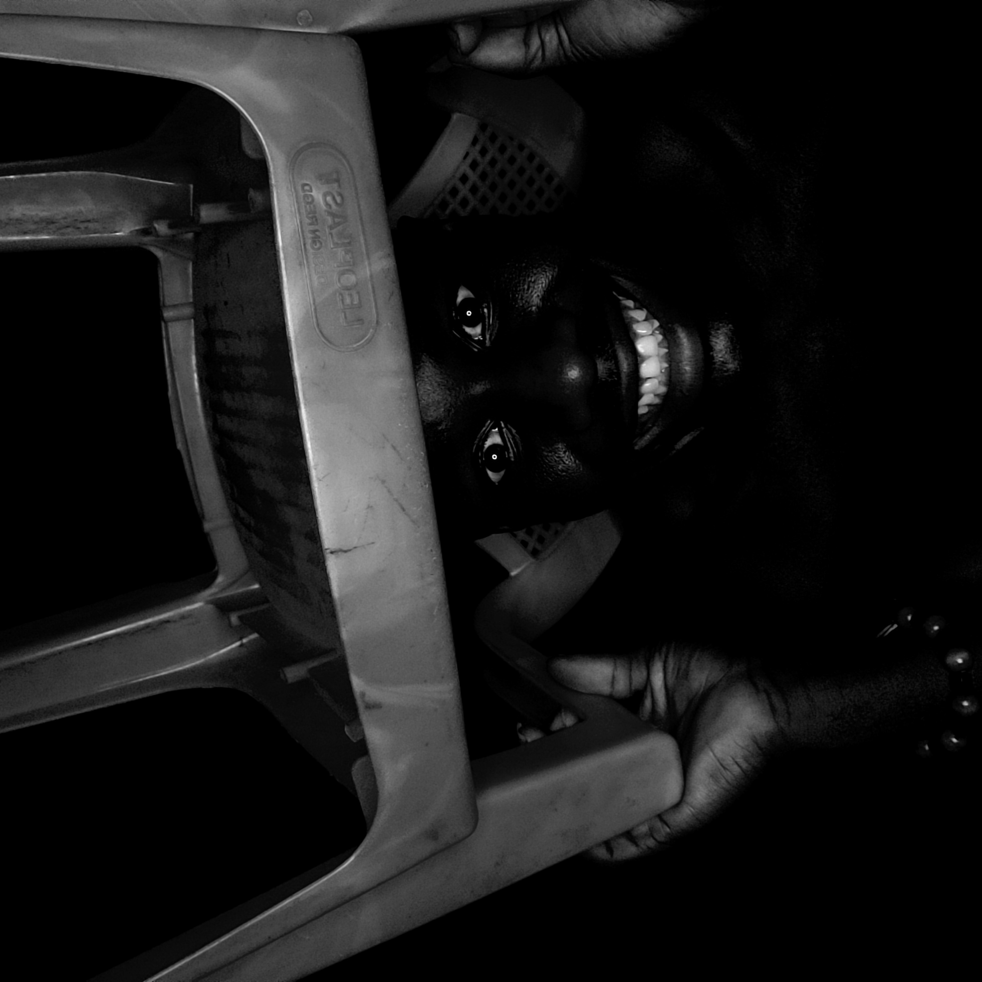 A high-contrast, black and white photograph captures a person peeking from under a light-colored plastic chair, against an almost entirely dark background. The image is oriented with the chair's frame running diagonally across the left side, and the person's face appearing on the right.
The person, with dark skin, exhibits a wide, radiant smile, showcasing a full set of bright white teeth. Their eyes are open and appear luminous, reflecting light. The overall expression conveys joy, playfulness, or even a mischievous quality, enhanced by the unusual perspective. Two hands are visible, gripping parts of the chair's frame, indicating the person is actively holding or interacting with the chair.
The chair is a common plastic stacking type, light in color and showing some signs of wear. On the chair's frame, mirrored text is visible, reading "GEORGES" and "DESIGN BY LEED". The surrounding environment is submerged in deep shadow, emphasizing the illuminated face and chair. The photograph creates an intimate and striking portrait, set in a low-light environment in Narayi, Nigeria.
