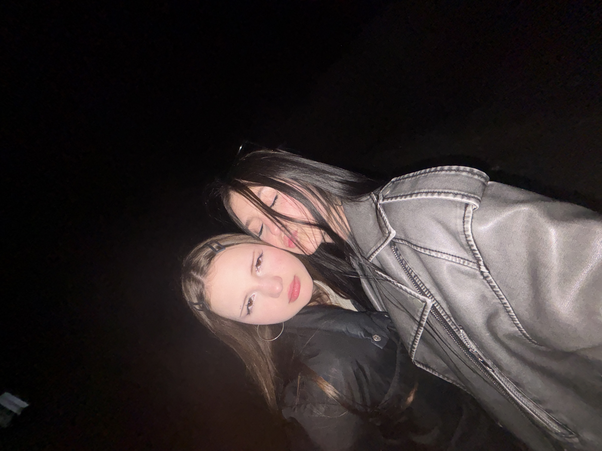 Two young women are closely posed together outdoors at night. One woman with long, dark hair wears a gray, weathered leather jacket and her eyes are closed. The other woman, with long, light-brown hair, wears a black jacket and silver hoop earrings. They are standing in front of a dark background. The location in the city of Фоки, Russia, cannot be determined from the image.