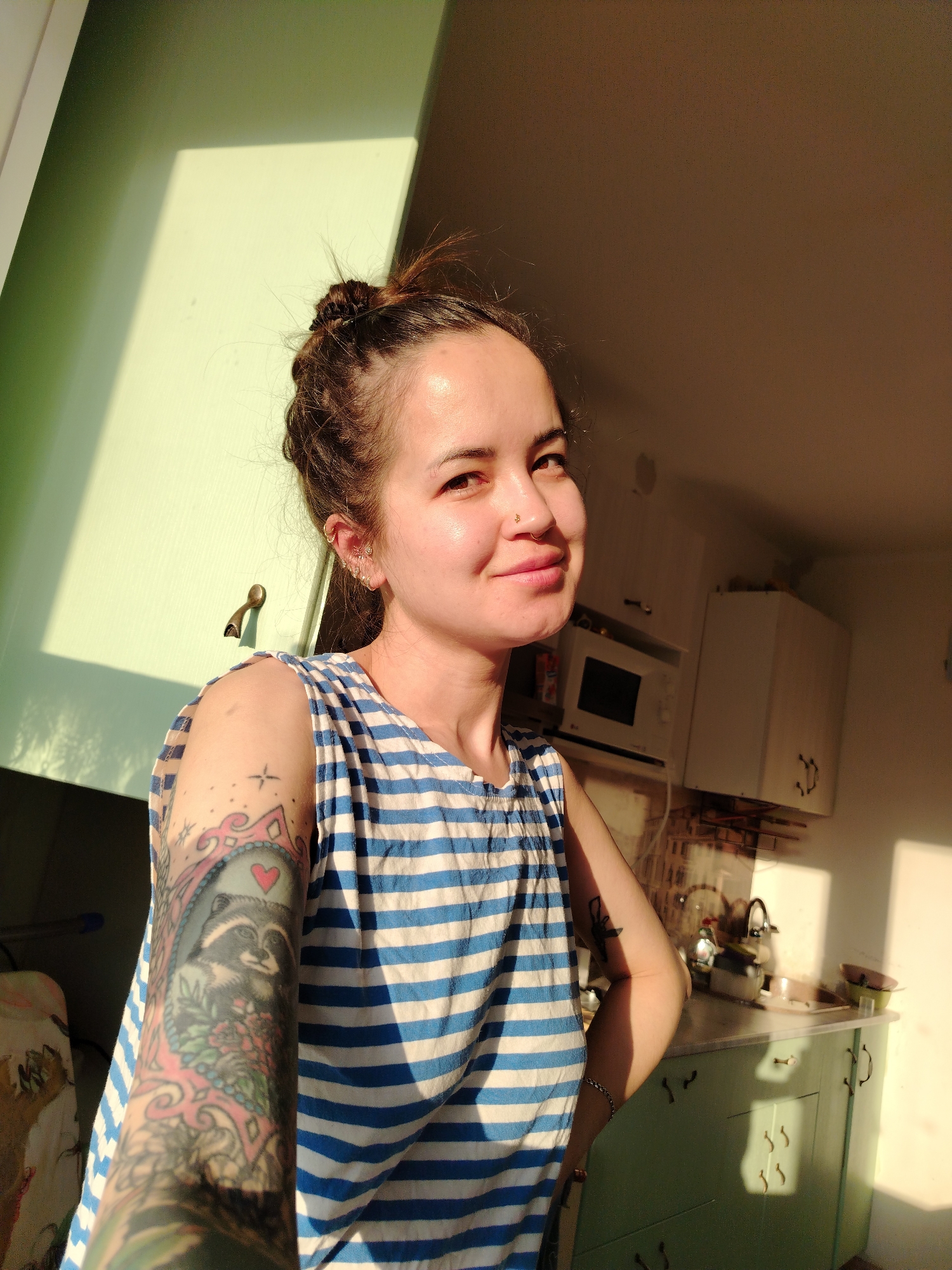 A fair-skinned woman with brown hair styled in a messy bun is smiling towards the camera. She wears a blue and white striped tank top. A full-sleeve tattoo is visible on her left arm. She has a nose piercing and multiple earrings in her left ear. The scene is indoors, likely a kitchen, with a microwave, cabinets, and a sink visible in the background. Sunlight illuminates the scene. There is no specific evidence the photo was taken in Noviy, Russia.