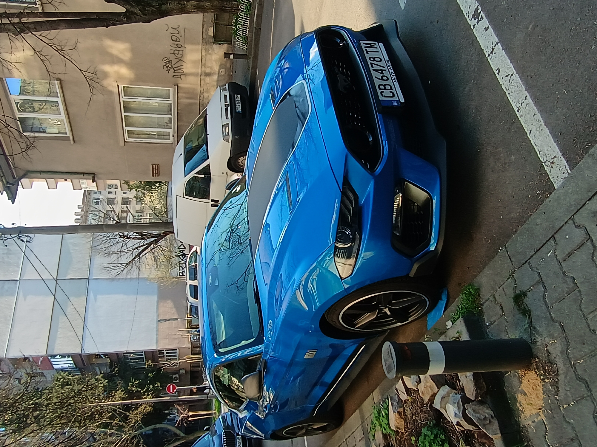 The image shows a blue Ford Mustang parked on a street in Stara Zagora, Bulgaria. The car has a Bulgarian license plate "CB 6478 TM." A white van is parked behind the Mustang. The street is lined with a building on one side. The building has graffiti. A street sign is visible. The weather appears to be sunny. A black and white bollard is close to the side of the car.