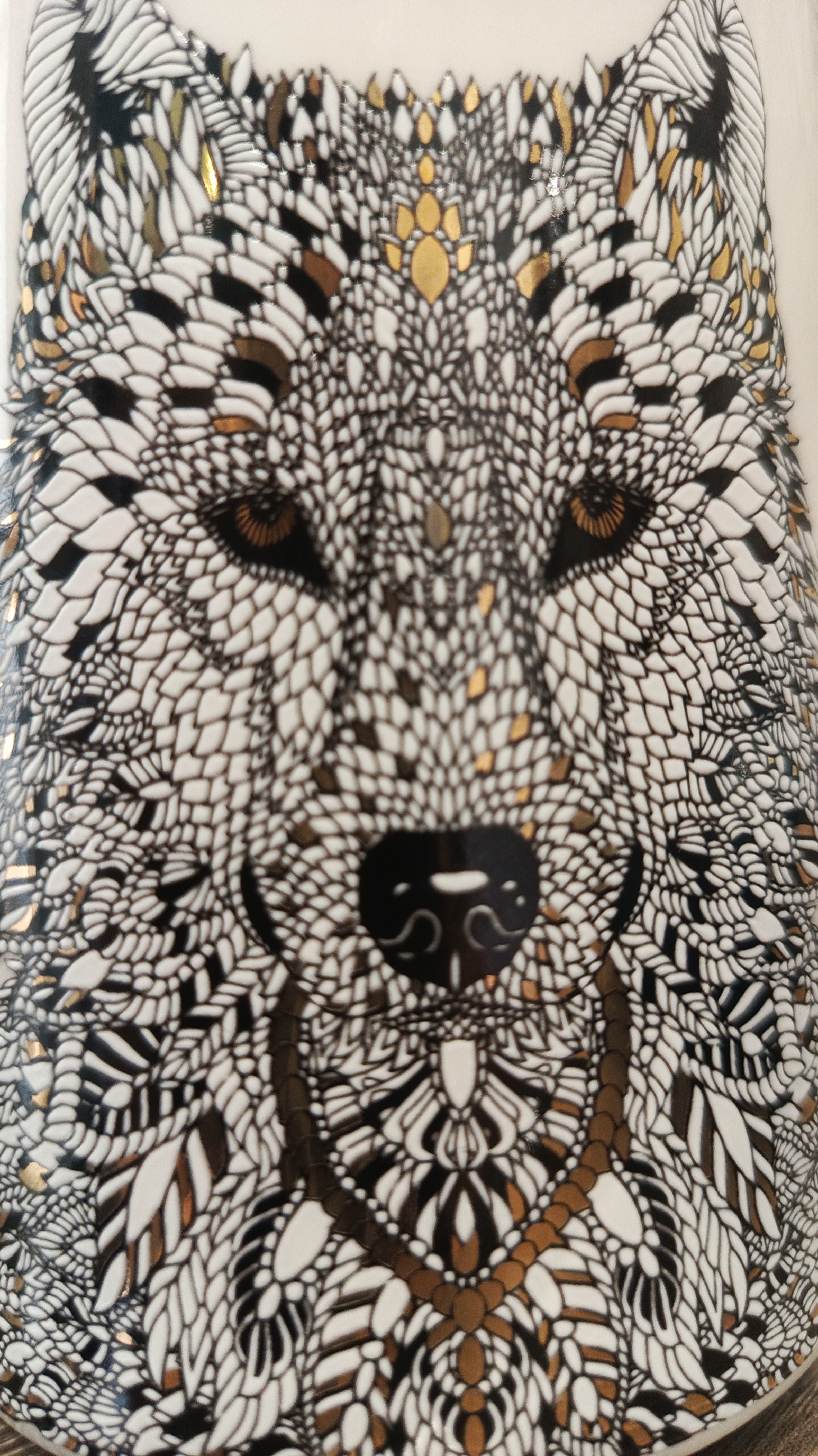 The image is a close-up, top-down view of a white ceramic object, likely a mug or vase, decorated with an intricate black, white, and gold pattern that forms the face of a wolf. The wolf's eyes are a warm brown, with a dark pupil. Its nose is black and heart-shaped, with a lighter colored detail on the bridge. A decorative collar-like band, composed of interconnected gold and black shapes, encircles the wolf's neck area. The fur is depicted with a repeating scale-like pattern, with some scales highlighted in gold and black. The lighting suggests an indoor setting, and the object appears to be placed on a flat surface, though the surface itself is not clearly visible. There is no indication of the city of Zvenigorod, Russia, or any other location.