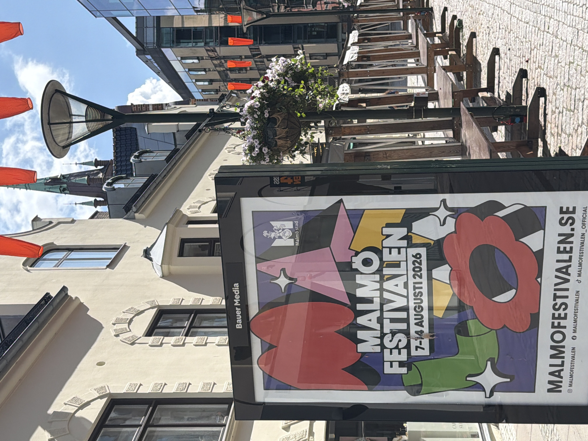 The image captures a street scene in Malmö, Sweden, featuring a poster for the "Malmöfestivalen" (Malmö Festival). The poster is prominently displayed on a black stand in the foreground, showcasing colorful abstract shapes and text that reads "MALMÖ FESTIVALEN" and "7-14 AUGUSTI 2026". Below this, "MALMOFESTIVALEN.SE" is visible, along with social media icons for Instagram, Facebook, and TikTok.
Behind the poster, the street is lined with outdoor seating arrangements, possibly for cafes or restaurants, consisting of wooden benches and tables. The street is paved with cobblestones. In the background, traditional European architecture with light-colored facades and arched windows is visible. A modern glass-fronted building also stands in the background, reflecting the sky and surrounding structures. Red decorative lanterns hang overhead, strung across the street. A lamppost with a hanging basket filled with flowers stands to the right of the poster. The sky is partly cloudy. No people are clearly visible in the scene. The overall impression is of a public urban space adorned for an event.