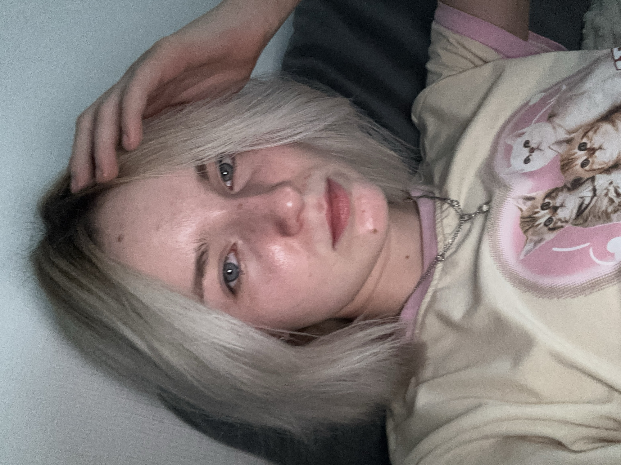 A fair-skinned young woman with blonde hair is reclining, possibly on a bed or sofa. She is wearing a light-colored top with a pink trim and a cat-themed graphic. Her left hand is resting on her head. She has light blue eyes and freckles. The background is out of focus, showing dark gray fabric and a white wall. The setting appears to be indoors. Location: Unknown, Russia.