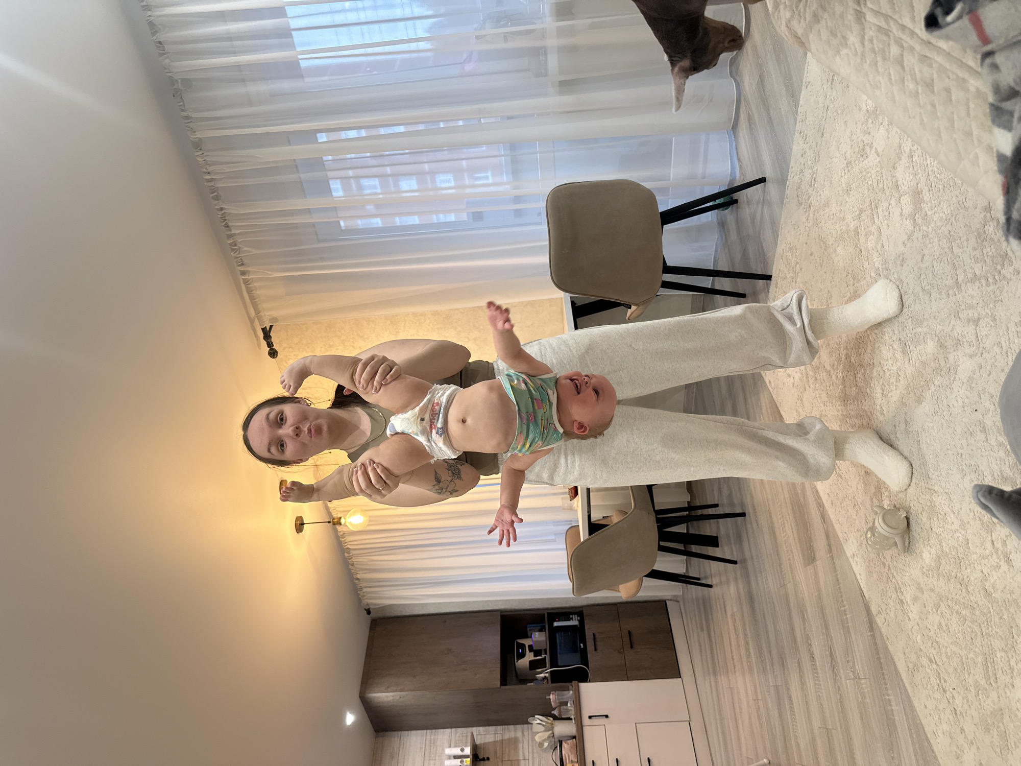 A woman lies on her back with a baby held above her, indoors, seemingly in a home in Chaikovsky, Russia. The woman wears light gray sweatpants and white socks. The baby wears a diaper and a patterned top. A dining table with two chairs is near the woman. The background includes a kitchen area with cabinets and appliances. A rug and a partial view of a sofa are also visible. Curtains cover a window in the background.
