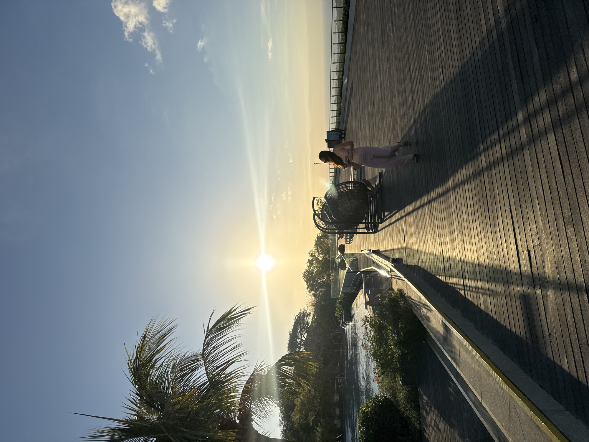 In Kuta Selatan, Indonesia, during a sunset, a woman in a light pink outfit walks along a wooden boardwalk. The sun, low on the horizon, casts long shadows across the boardwalk and illuminates the scene with a warm, golden light. To the left of the boardwalk, a swimming pool is visible, bordered by lush greenery. A palm tree extends into the upper left corner of the frame, its fronds creating a silhouette against the clear blue sky. In the distance, the ocean stretches out to meet the horizon. A large, circular fan-like structure stands on the boardwalk, casting a shadow. The woman appears to be holding a phone in her hand.