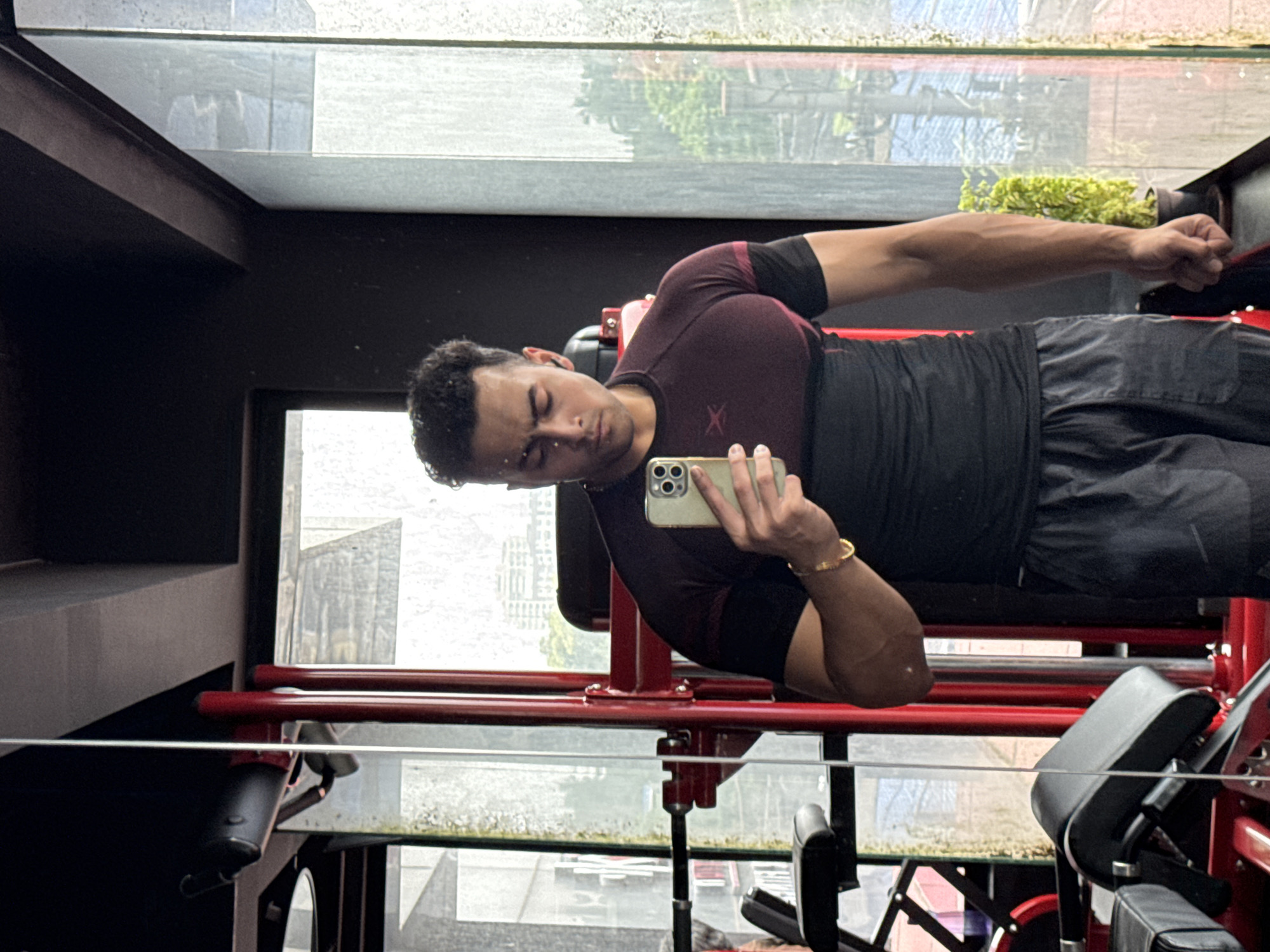 A man is taking a mirror selfie in a gym. He is wearing a maroon and black athletic shirt and black shorts. He has a gold bracelet on his left wrist. He is holding a smartphone in his right hand. The gym has red equipment and a large window with a view of the city. The view through the window shows buildings, trees, and a cloudy sky. It is likely raining outside. The man appears to be of Indian descent and is likely in his 20s or 30s. The overall atmosphere is that of a modern fitness facility. The image is taken from a medium-shot angle, capturing the man from the waist up.