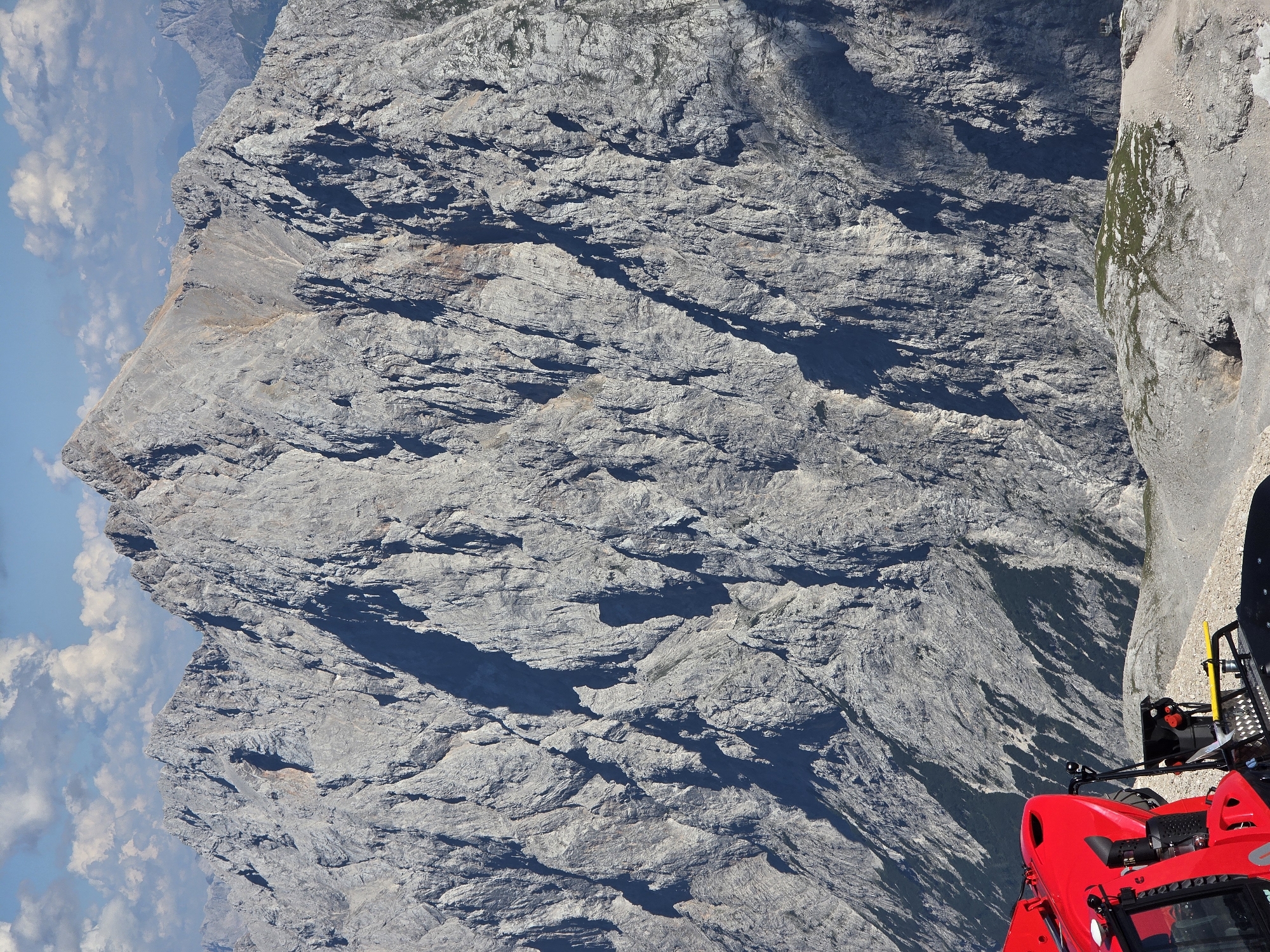 This is a photograph taken from a high vantage point overlooking a vast mountain range. In the foreground, a bright red snow groomer is partially visible, its mechanical components and sturdy frame suggesting it is used for maintaining snowy terrain. The machine is parked on a rocky, mountainous landscape. The dominant feature of the image is the imposing, rugged mountain face that fills the background, characterized by its sheer cliffs, deep ravines, and sharp peaks. The rock appears weathered and grey, with visible strata and textural variations. Patches of sparse vegetation are visible on some of the lower slopes. The sky above is a clear blue, dotted with fluffy white clouds, indicating fair weather. This scene is likely located in the Bavarian Alps, given the mention of Garmisch-Partenkirchen, Germany. Garmisch-Partenkirchen is known for its dramatic mountain scenery, including the Zugspitze, Germany's highest peak, which is consistent with the grandeur of the mountains depicted. The presence of the snow groomer suggests the image might have been taken during a season when snow is expected or has recently fallen, or it could be a permanent fixture at a high-altitude facility. There are no people visible in the image.